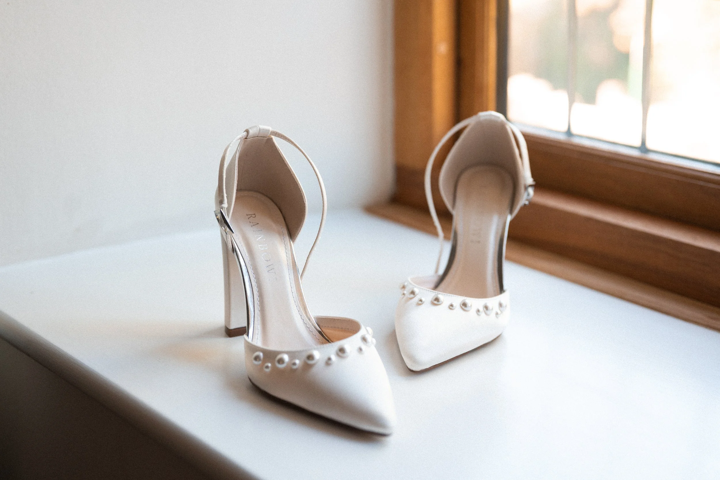 A pair of white high-heeled shoes with pearl accents on the toe, placed on a white surface near a wooden window frame.