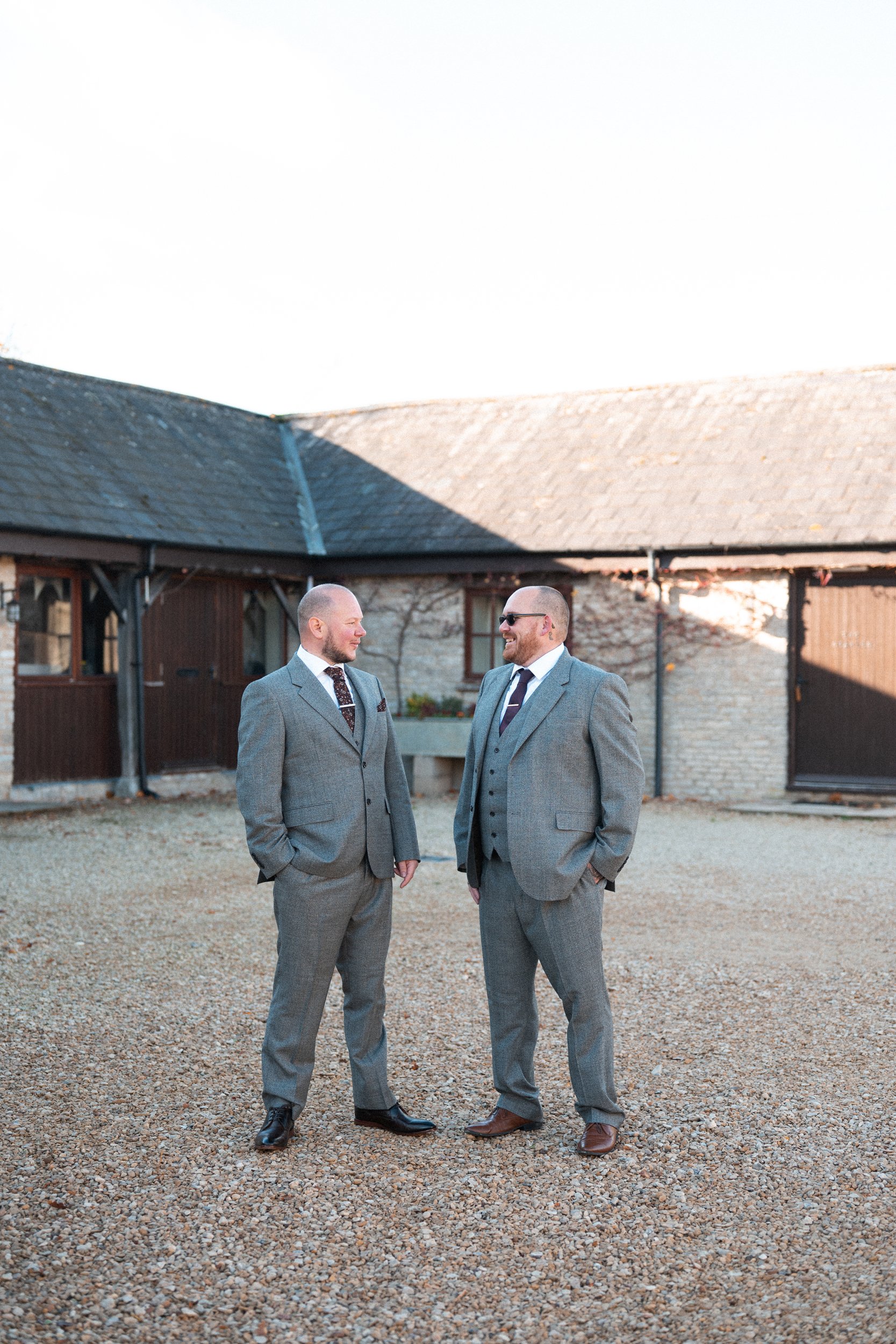 Two men in gray suits standing outdoors, smiling at each other, with a rustic building in the background.