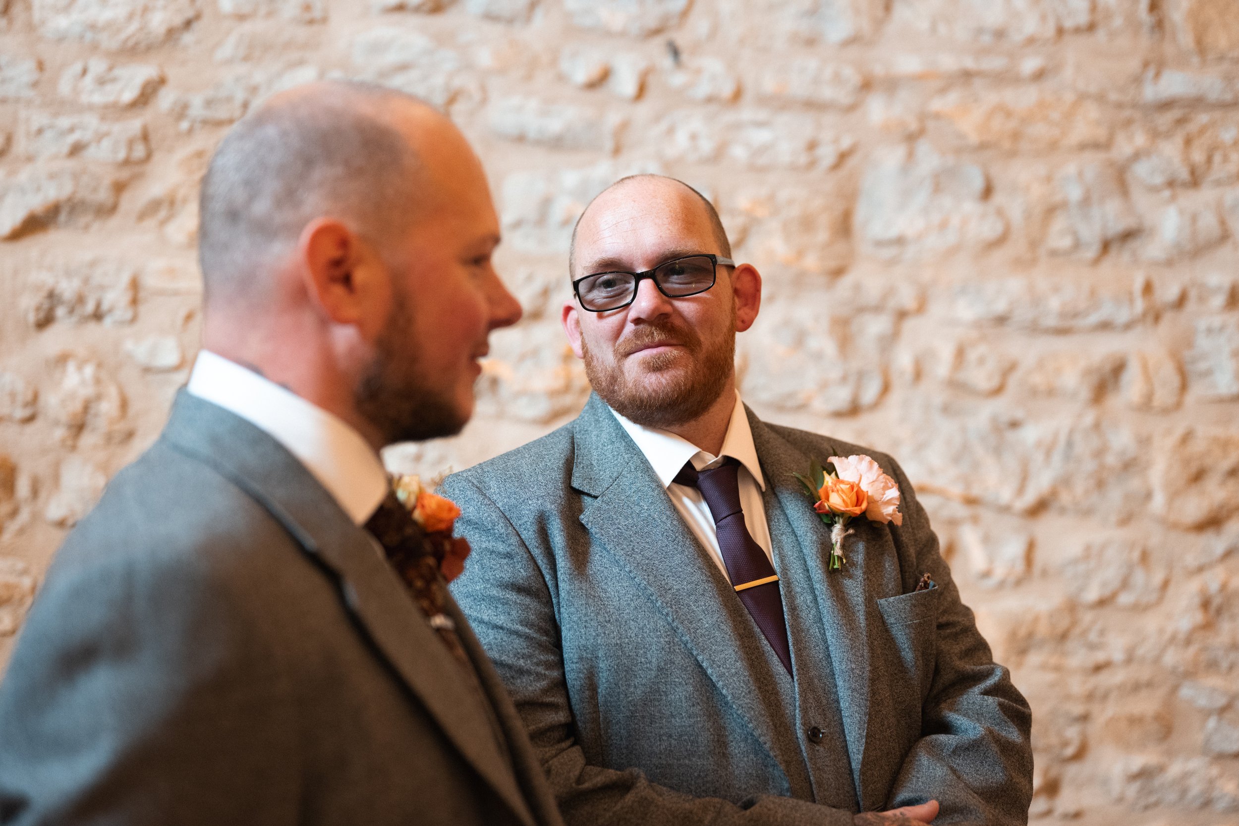 Two men in suits and glasses sharing a moment, one smiling softly, against a textured stone wall.