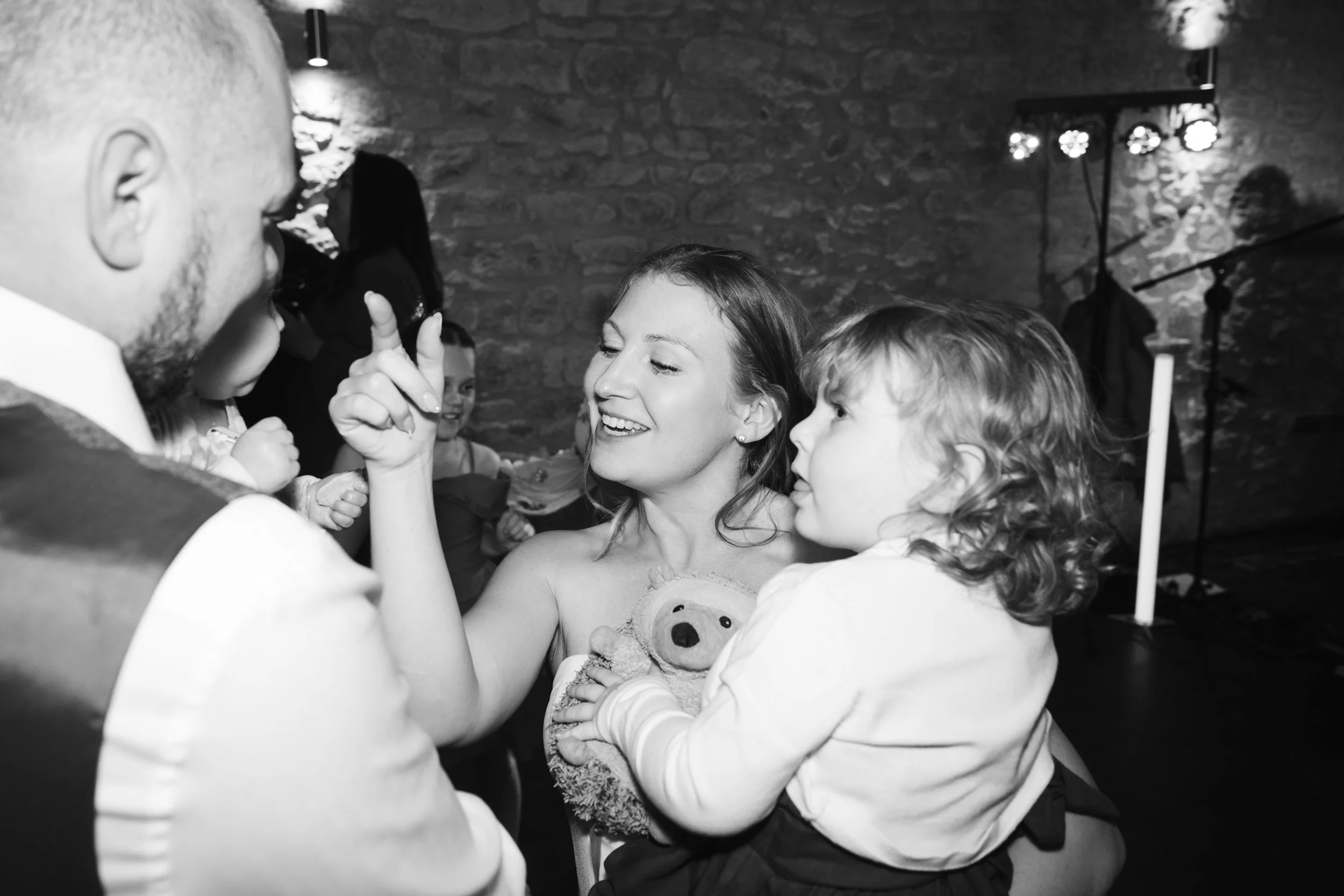 A woman and a young girl are looking at a man, with the woman smiling and holding the girl with a teddy bear. The woman is pointing with her finger at the man in an indoor setting with a brick wall background.