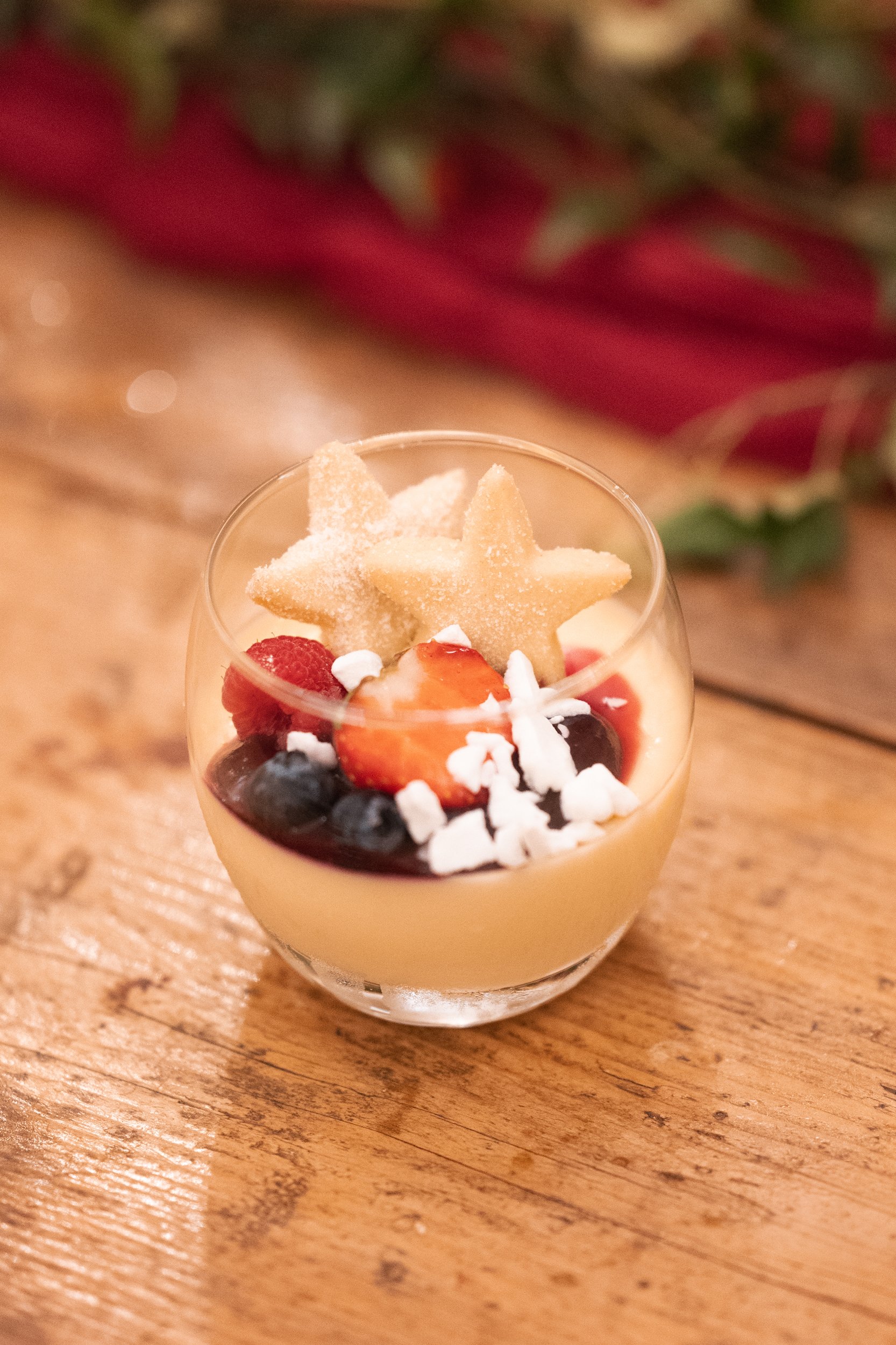 A dessert in a glass cup topped with star-shaped cookies, fresh strawberries, blueberries, white coconut flakes, and a strawberry sauce.
