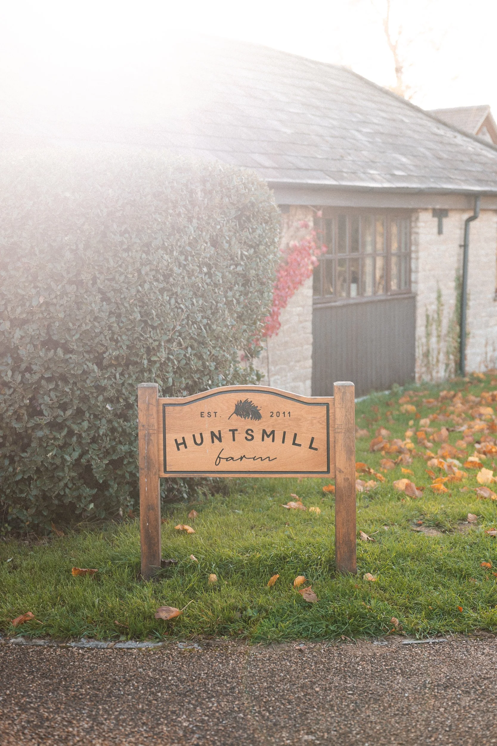 Wooden sign for Huntsmill Farm, established 2011, with a background of a stone building, bushes, and fallen leaves on the grass in an outdoor rural setting.