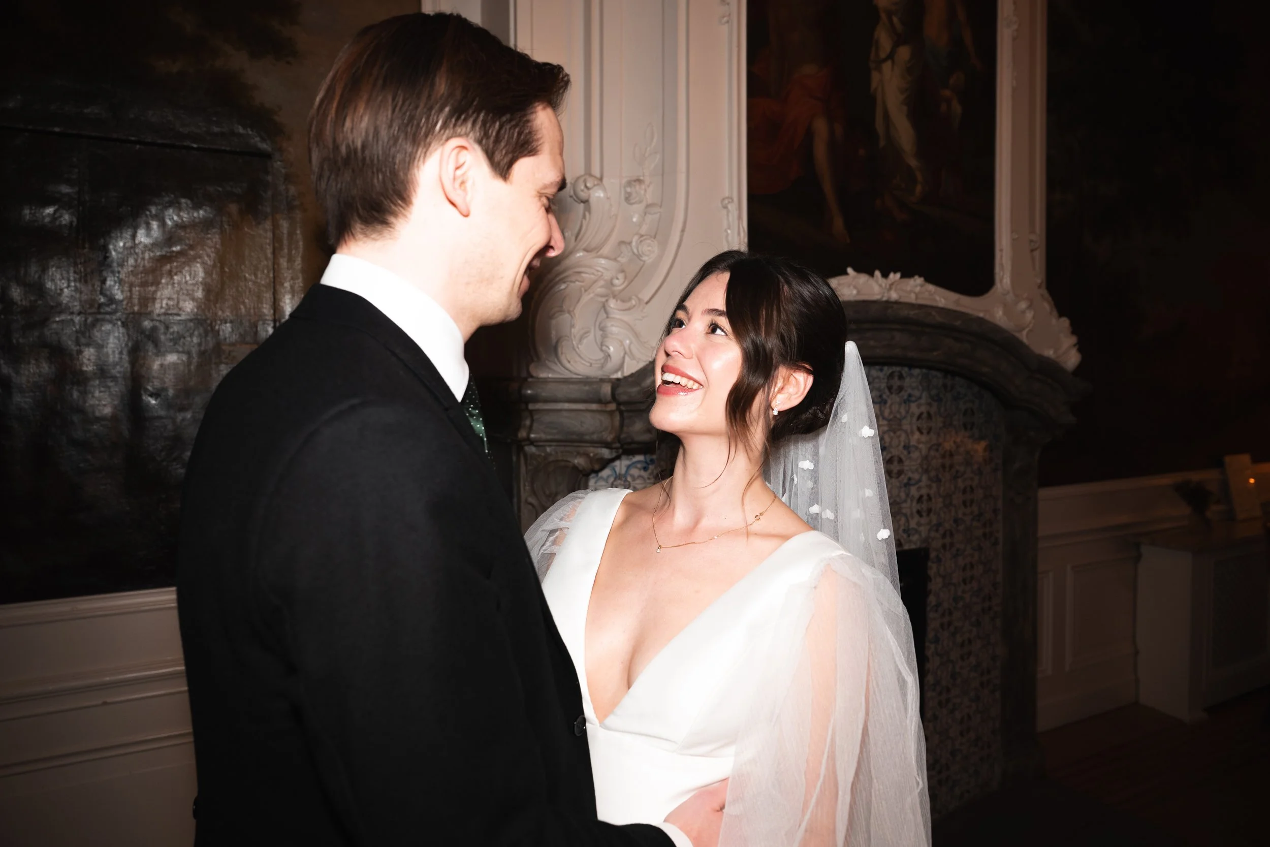 A bride and groom share a joyful moment at their wedding; the bride is looking at the groom with a bright smile, wearing a white wedding dress and veil, while the groom, dressed in a black suit, smiles back.