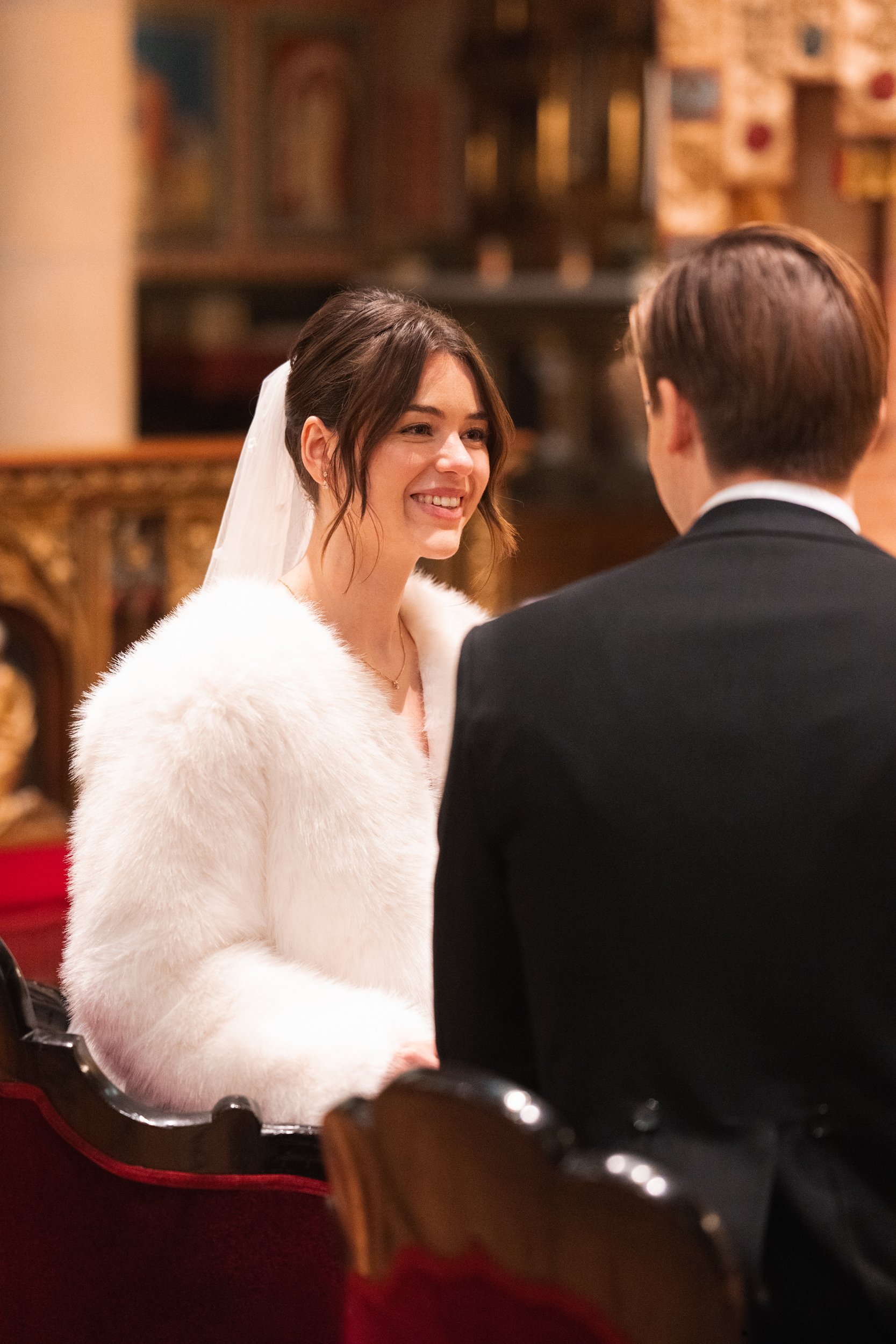 A bride with a white fur coat and a veil is smiling at a groom in a black tuxedo during a wedding ceremony inside a decorated venue.