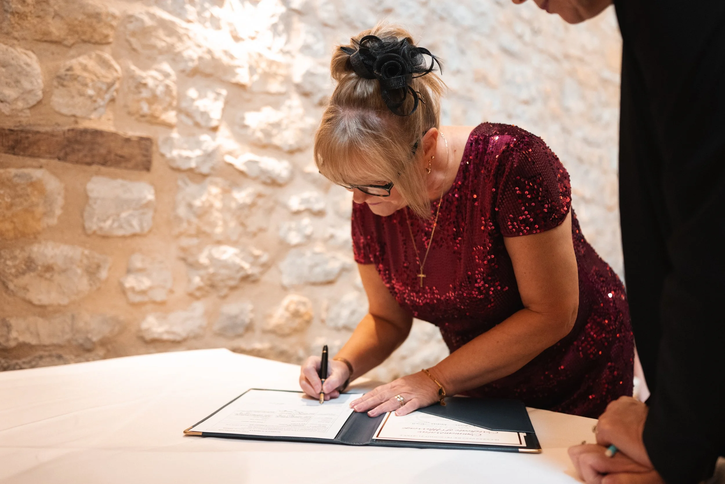 A woman in a red sequined dress with glasses and a black hair accessory signs a document on a table, with a man partially visible beside her.