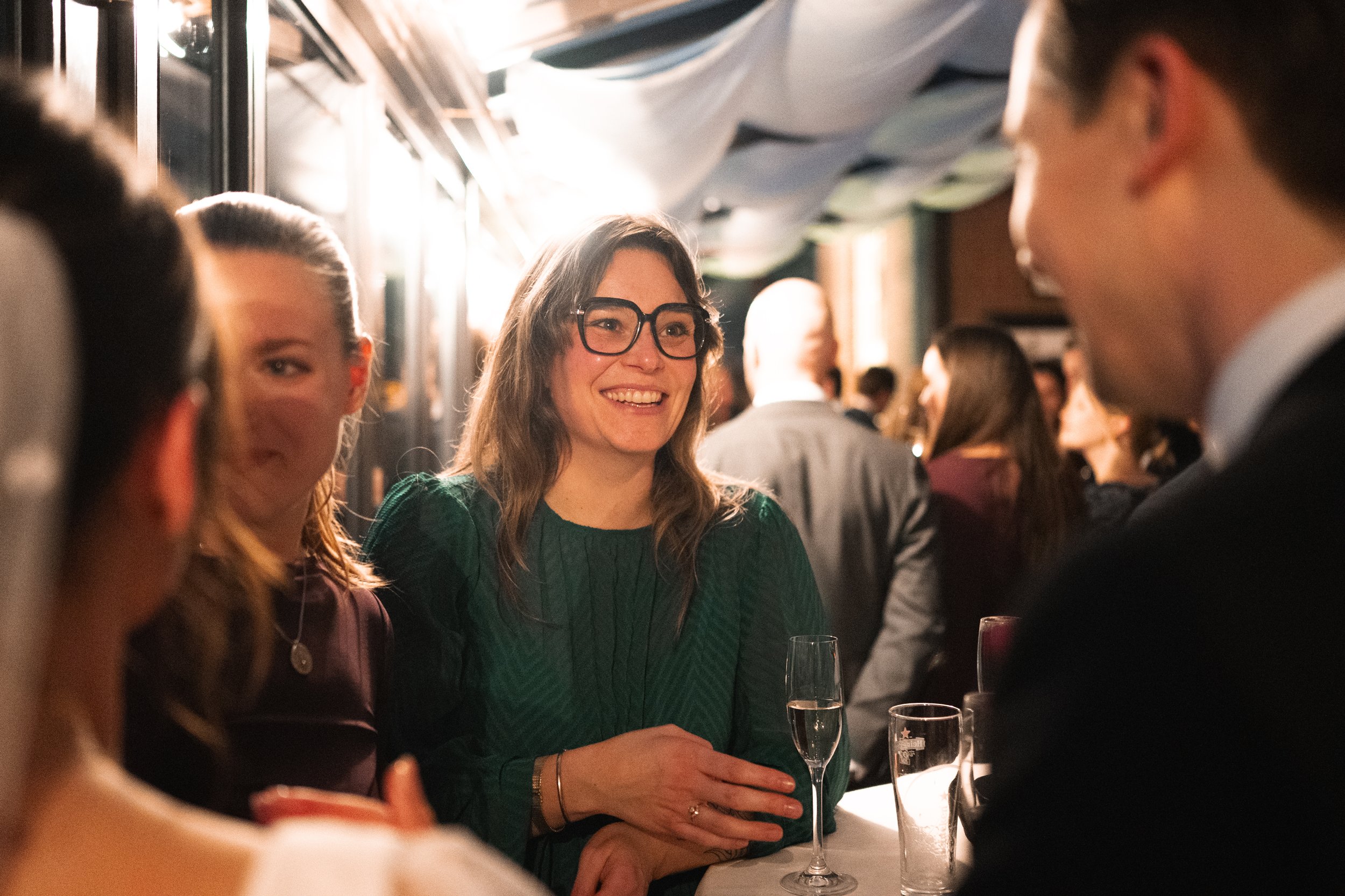 A woman with glasses smiling at a social gathering, surrounded by people and drinks in a dimly lit room.