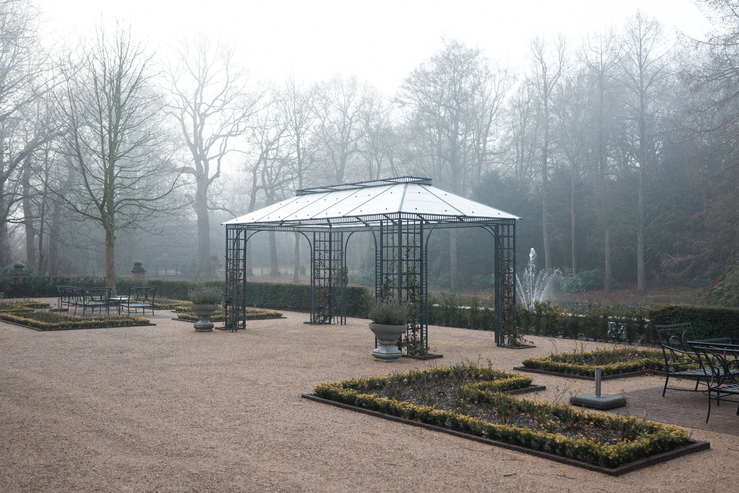 Empty garden with a metal gazebo, benches, and flower beds, surrounded by bare trees on a foggy day.
