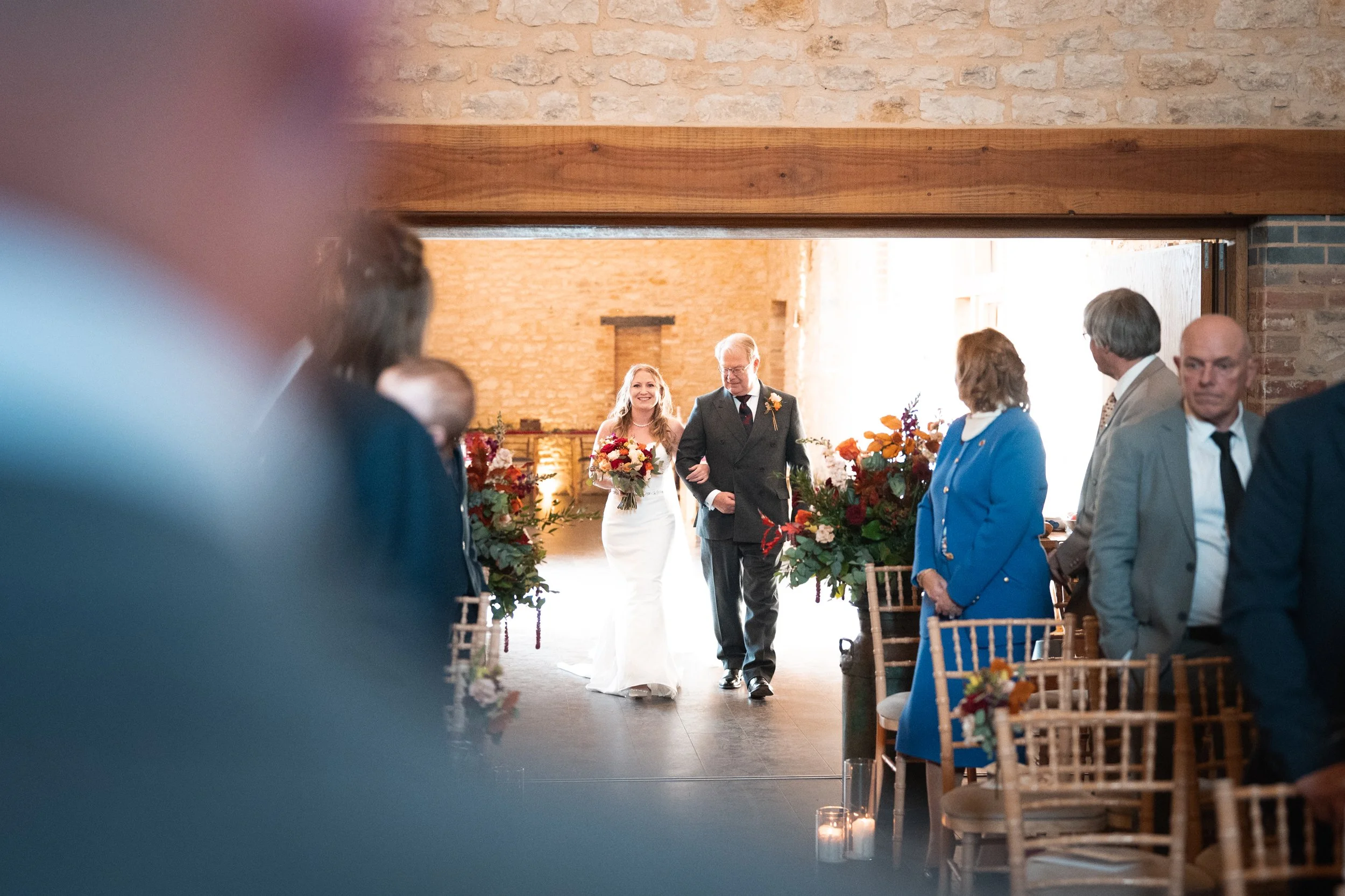A bride walking down the aisle with her father at a wedding ceremony inside a rustic venue with brick walls and wooden accents, surrounded by seated guests.