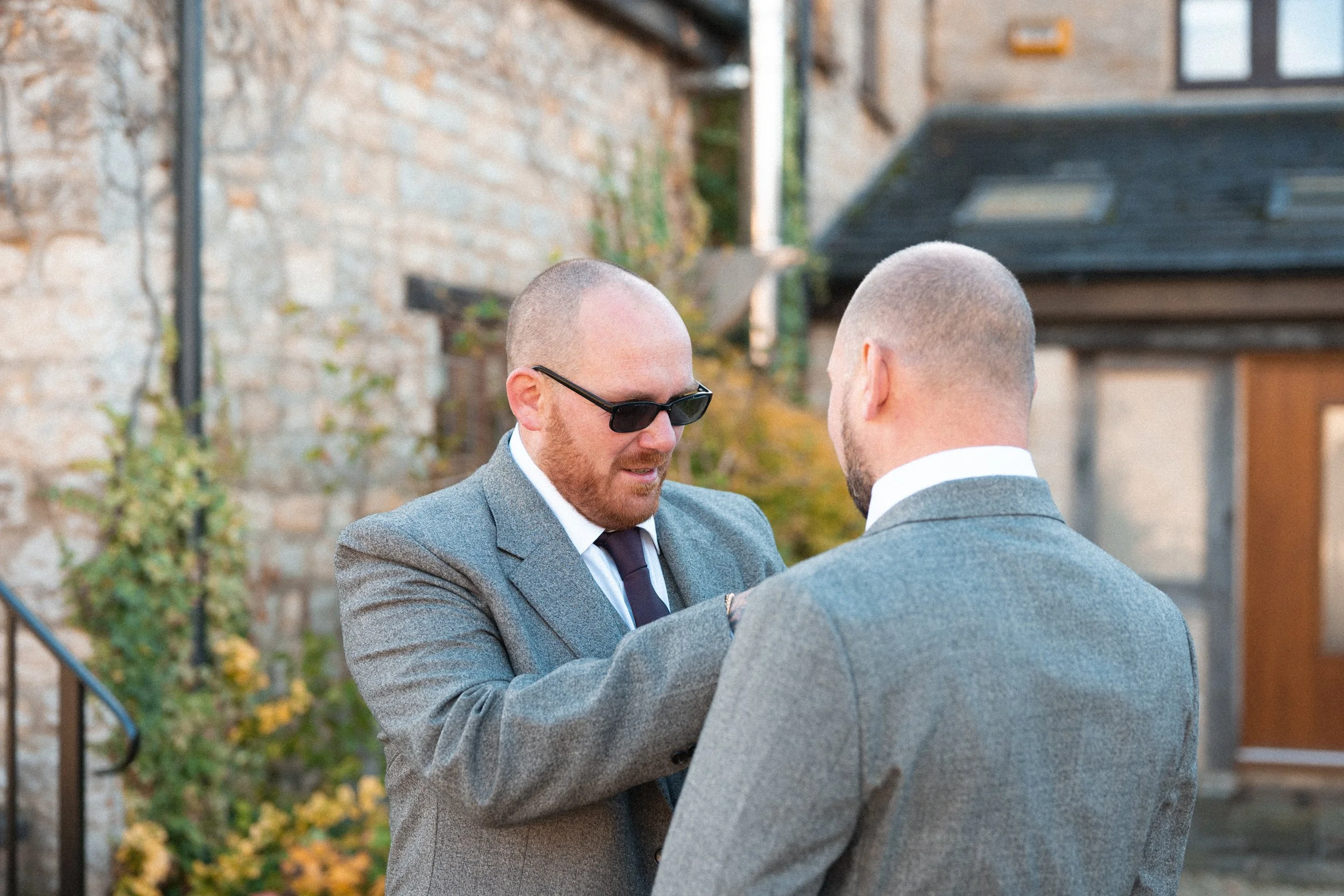 Two men in gray suits, one with sunglasses, talking outdoors in front of a brick building.