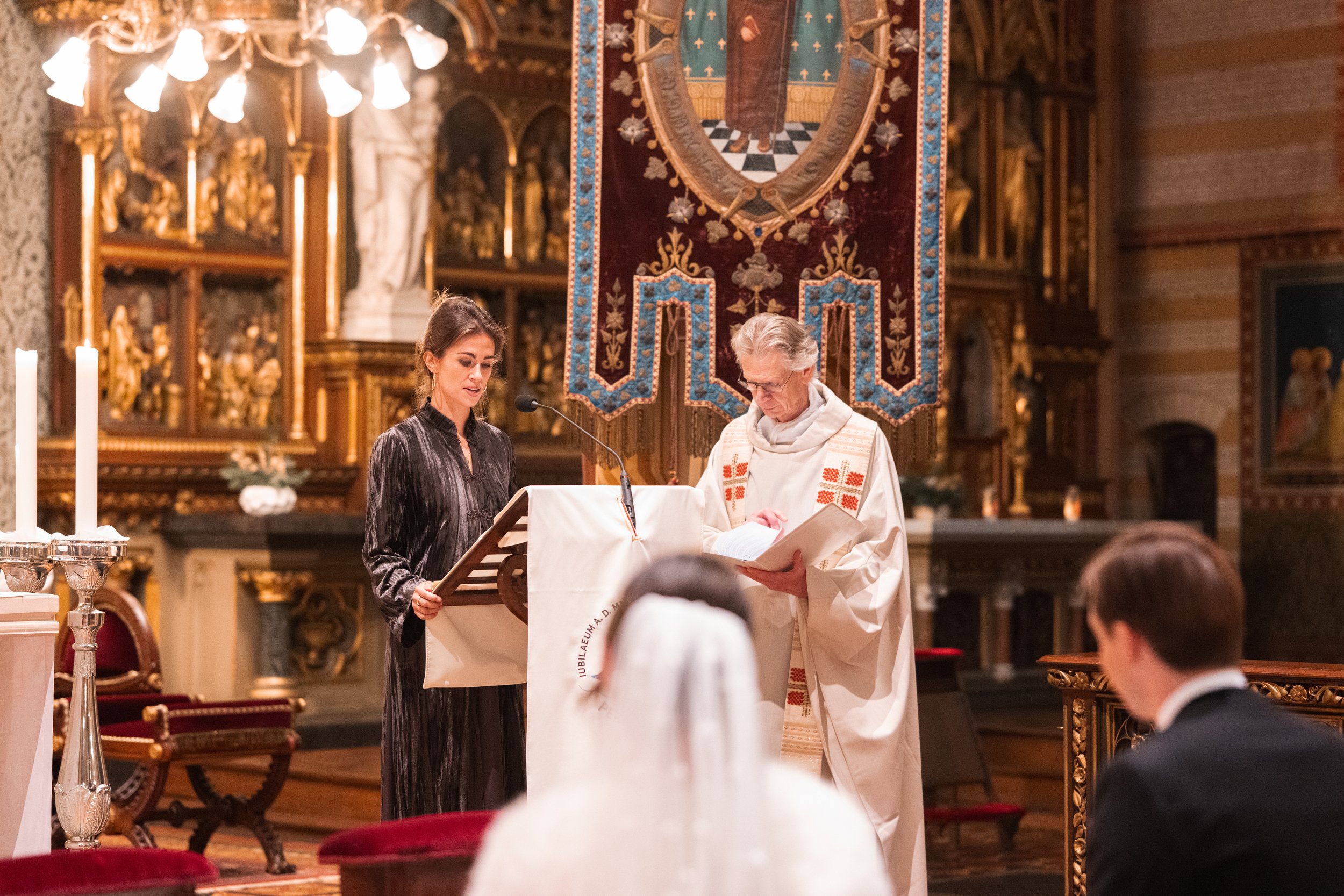 A woman and a priest standing at the altar of a church during a religious ceremony, with wood and gold decor, a large priest's cloth with religious symbols, and a large tapestry hanging behind.