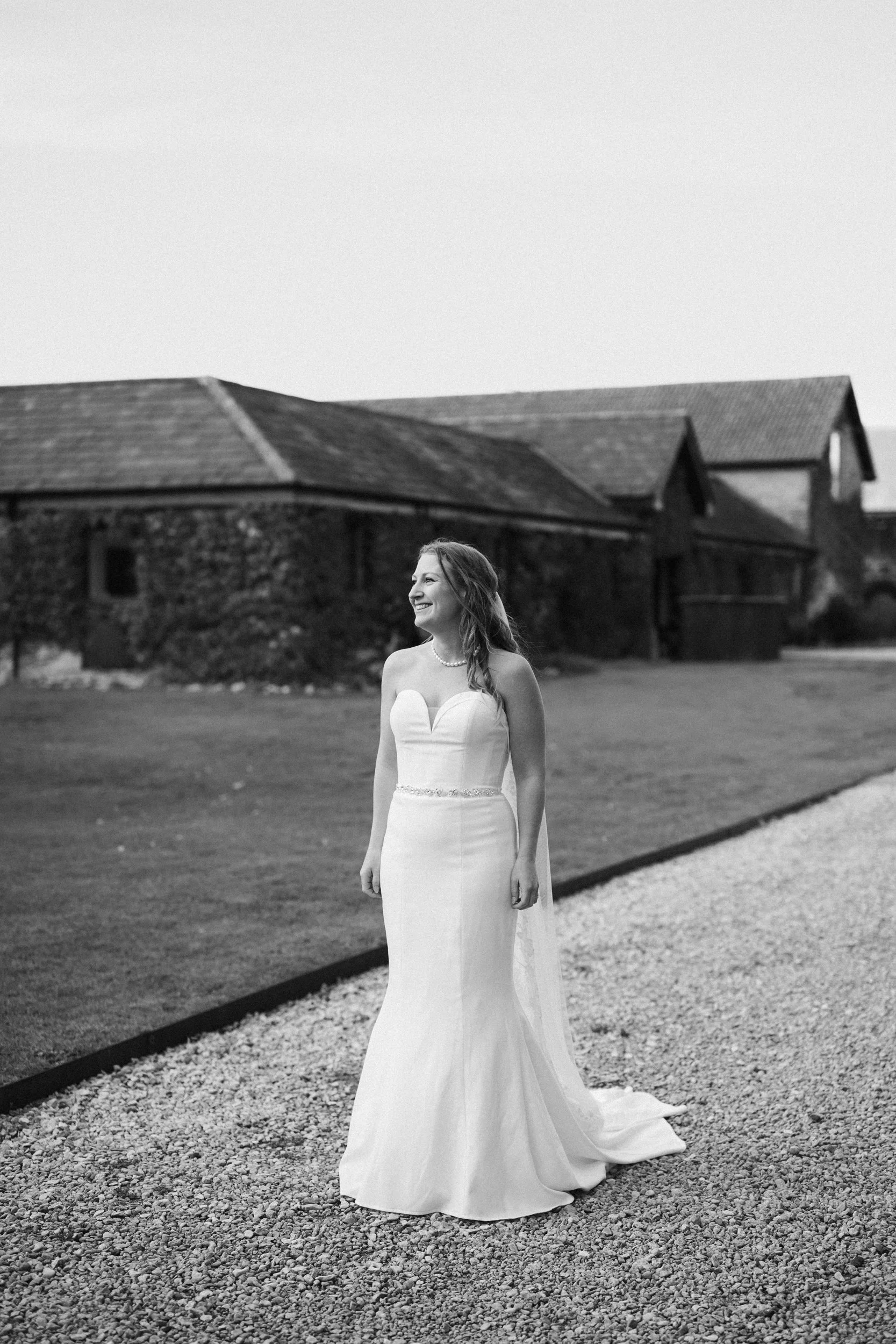 A woman in a wedding dress standing outdoors on a gravel path with a brick building and grass in the background.