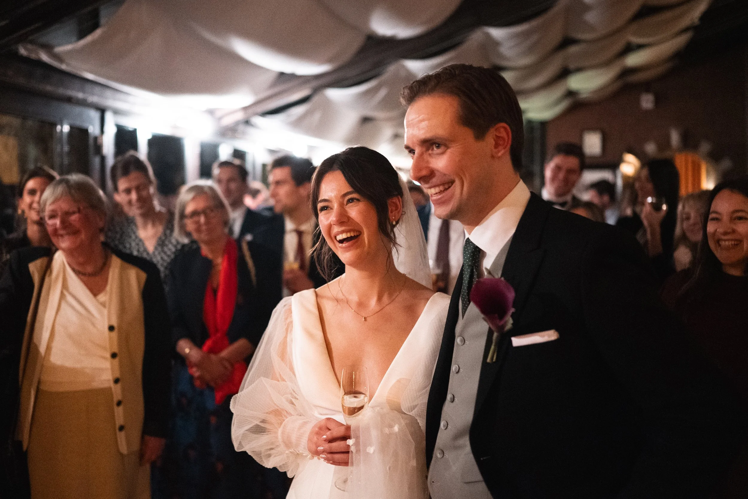 A bride and groom smiling at each other during their wedding reception, surrounded by happy guests.