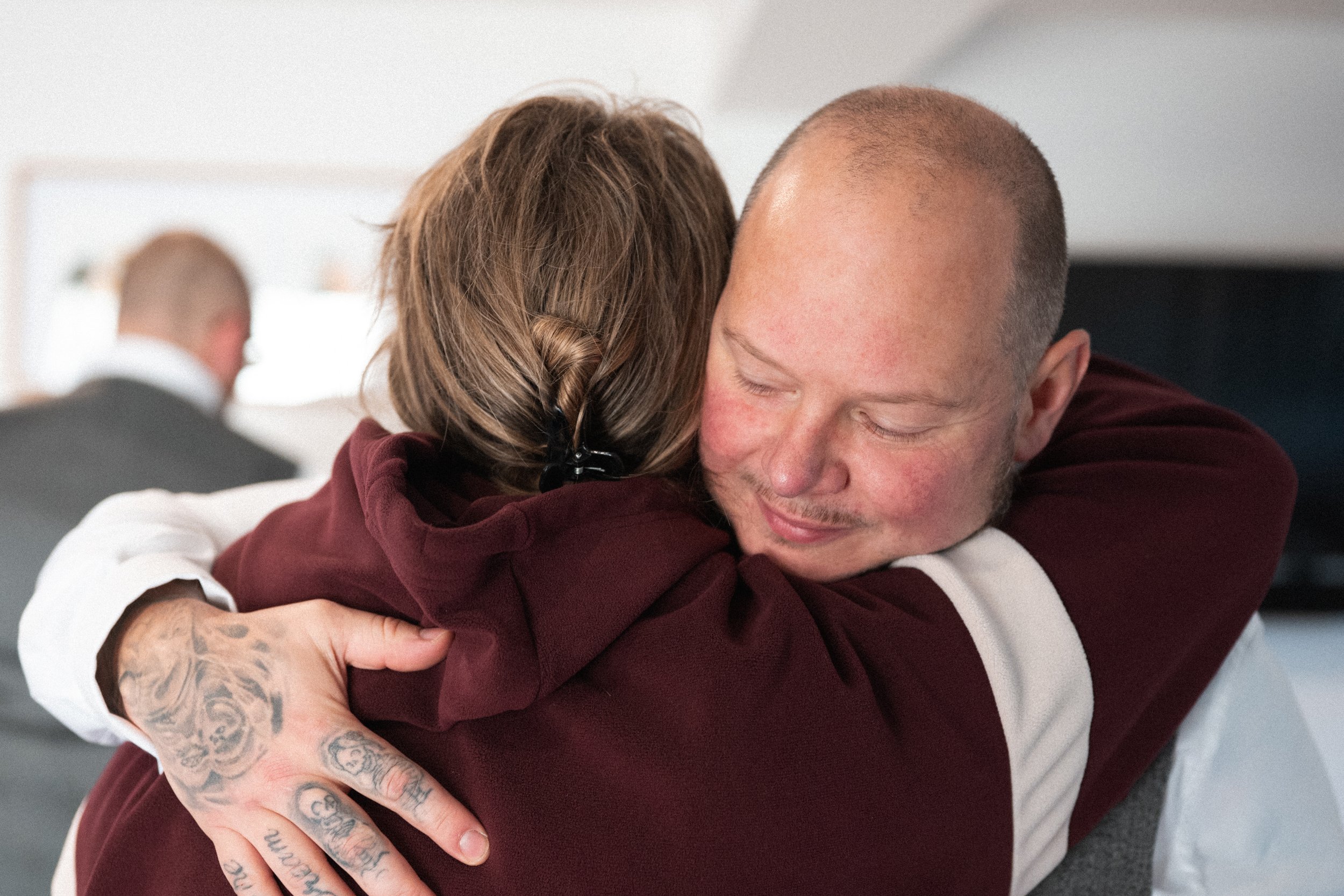 A man with a shaved head and tattoos on his hand hugs a woman with brown hair in a burgundy hoodie, smiling softly with eyes closed.