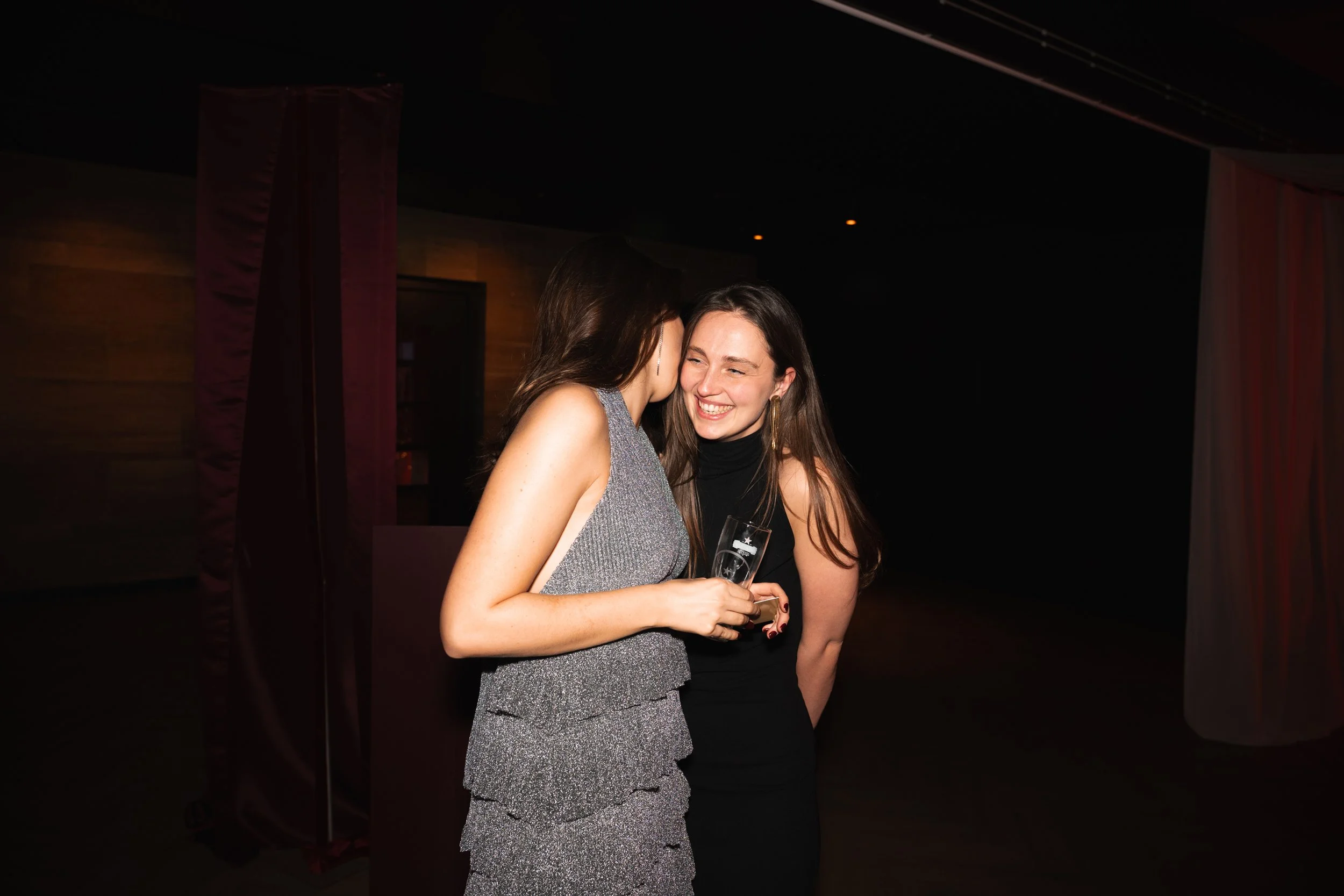 Two women at a social event, one whispering in the other's ear, holding a glass of champagne, smiling.