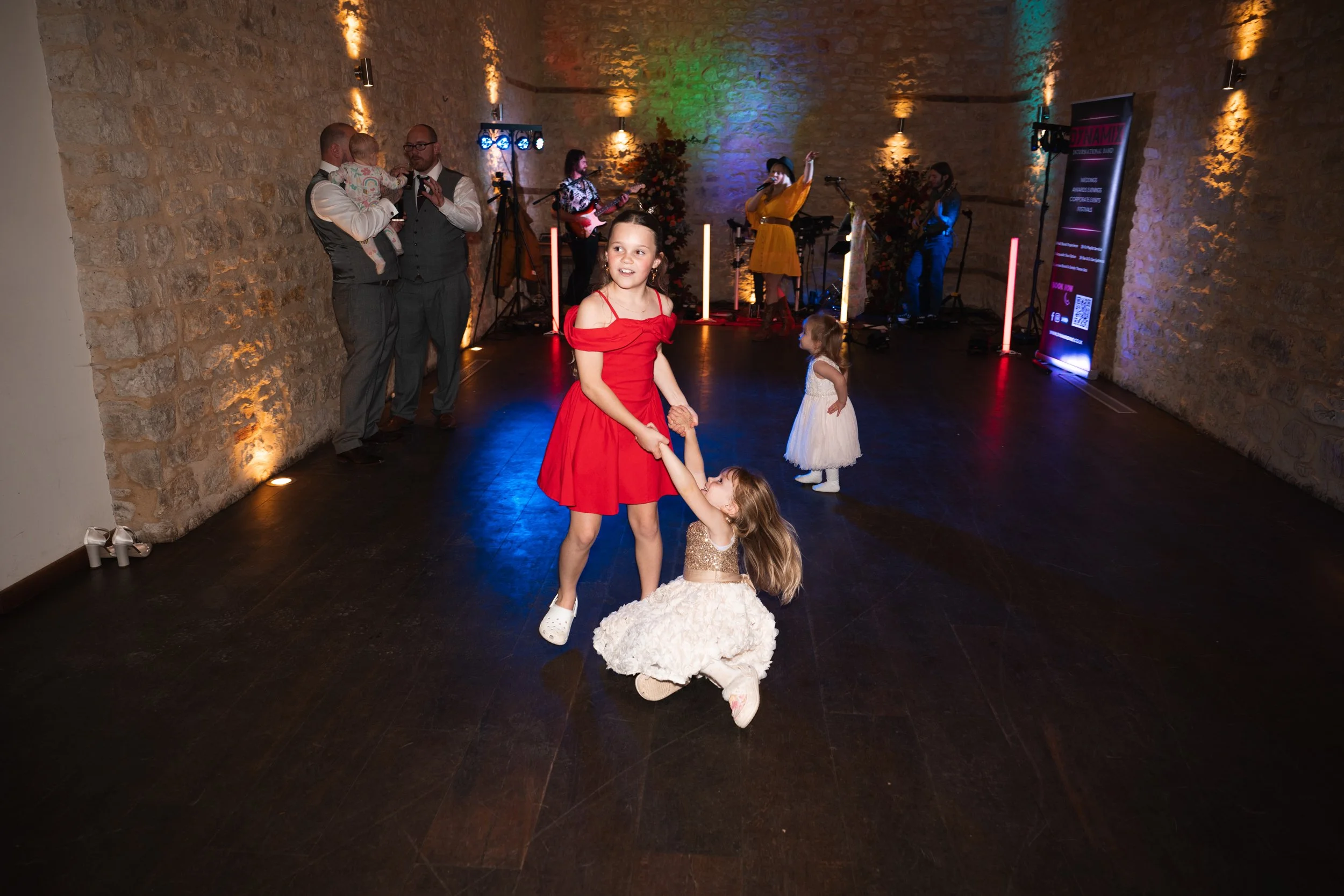 Children dancing and a band performing at a celebration or party in an indoor venue with stone walls and colorful lighting.
