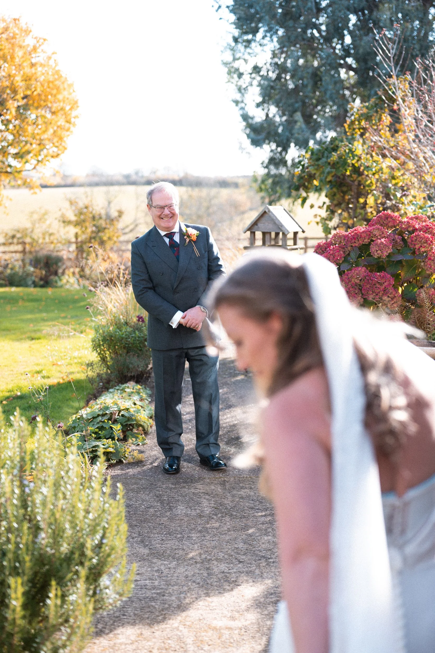 A groom in a dark suit standing outdoors, smiling while watching a bride in a white dress, with colorful flowers and trees in the background on a sunny day.