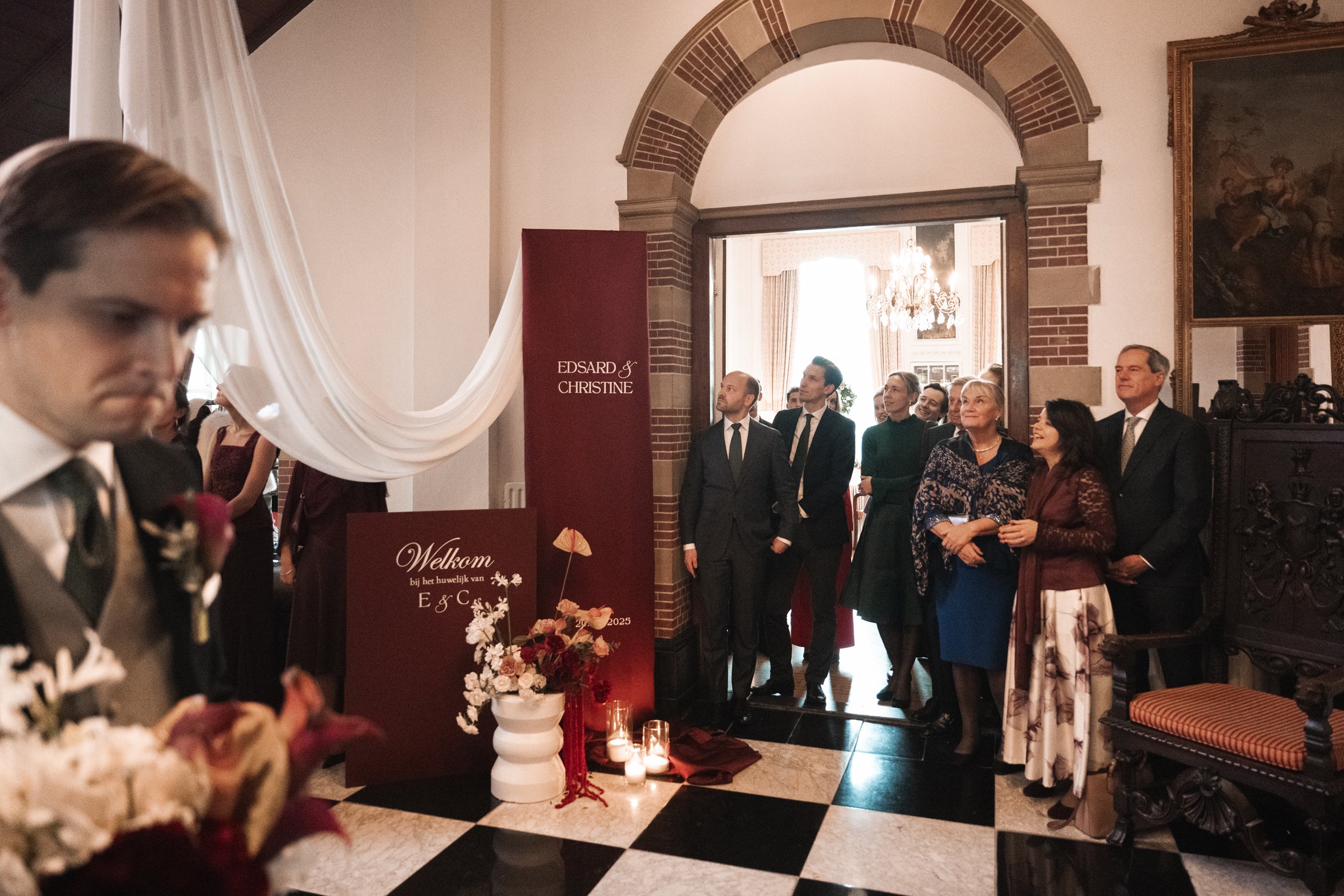 People gathered at a wedding entrance, with a sign saying 'Welkom bij het huwelijk van Edsard & Christine,' decorated with flowers and candles, inside a grand hall with high ceilings, chandeliers, and a painting on the wall.