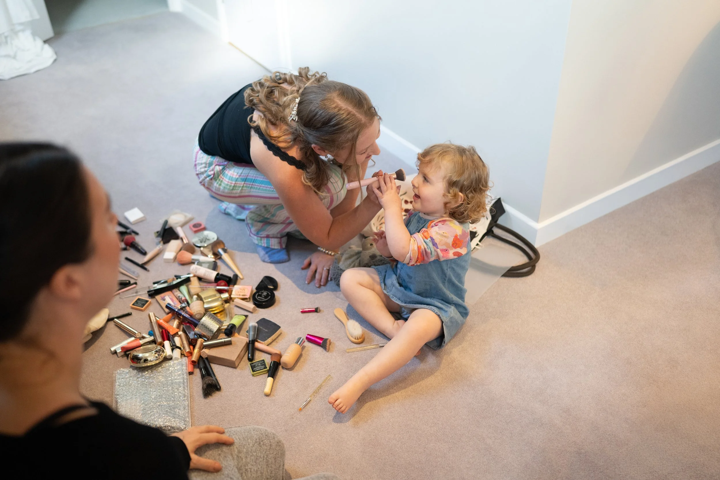 A woman applying makeup to a young girl sitting on the floor surrounded by makeup products, with another woman sitting nearby watching.