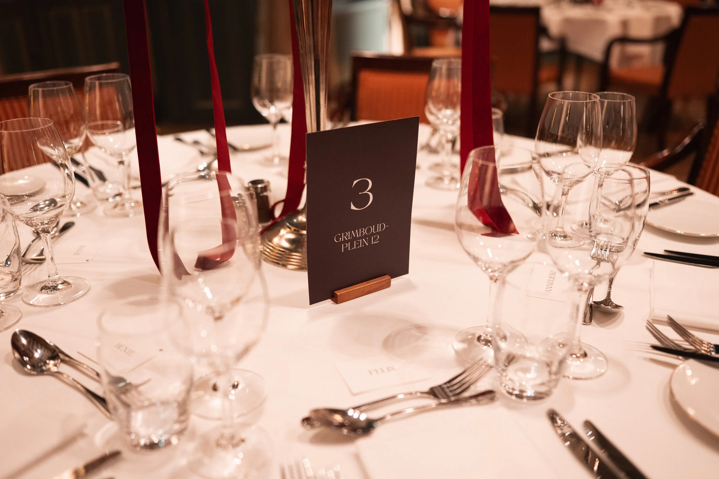 Formal dining table set with multiple wine glasses, silverware, a black table number sign displaying the number 3, and a dark red ribbon centerpiece.