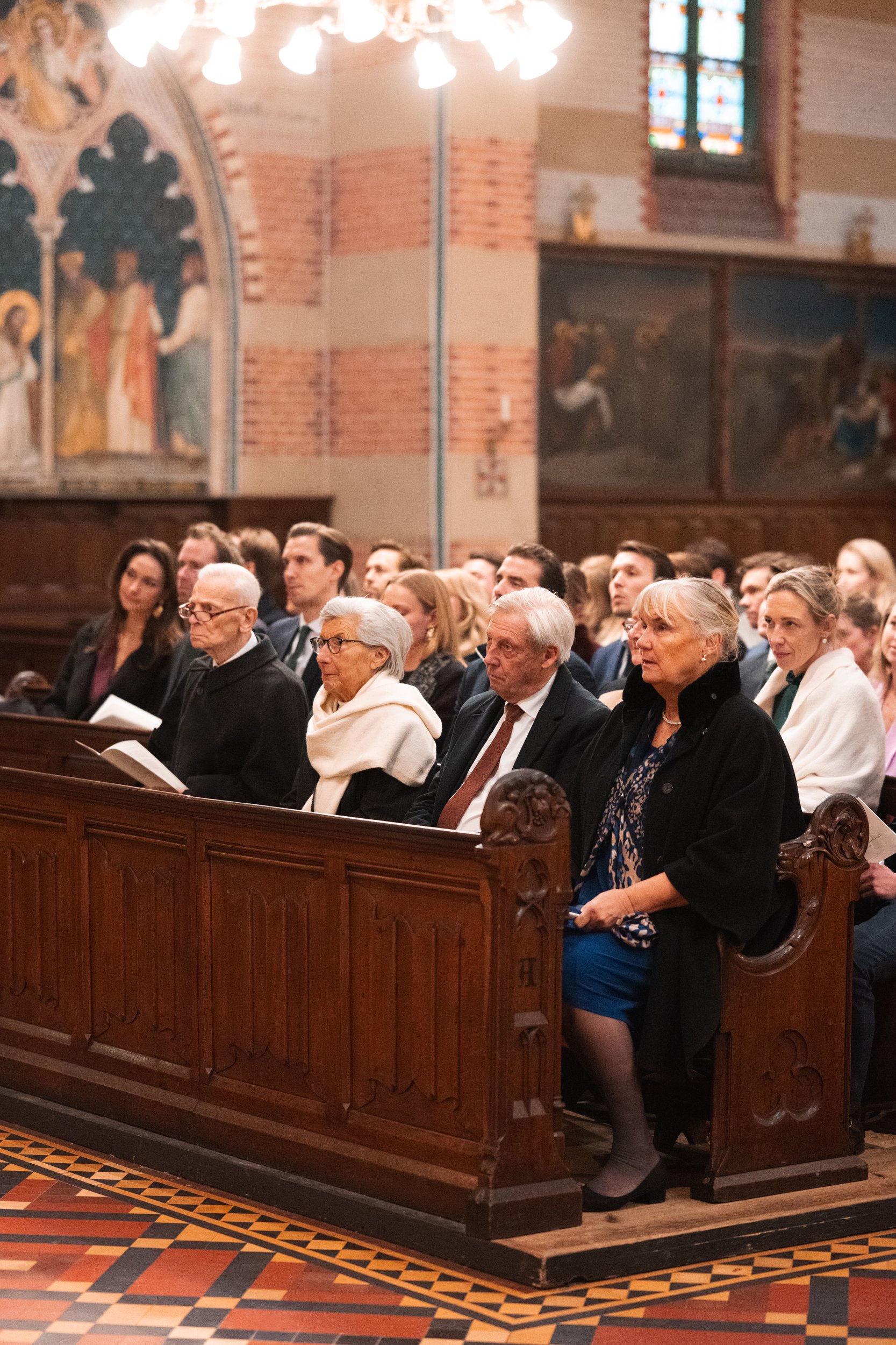 A group of people seated in wooden pews inside a church, with stained glass windows and religious artwork on the walls.