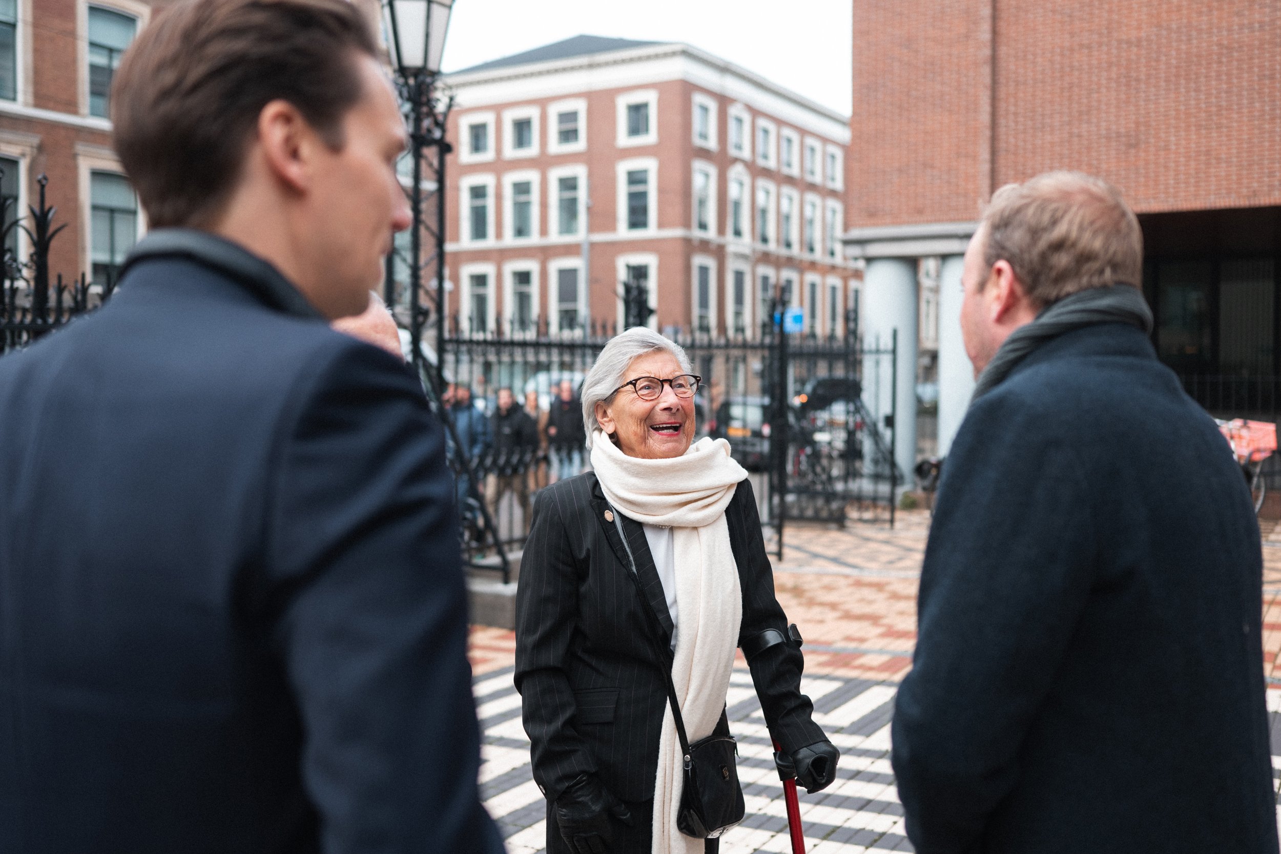 An elderly woman smiling and talking to two men on the street in front of a black iron fence and brick building.