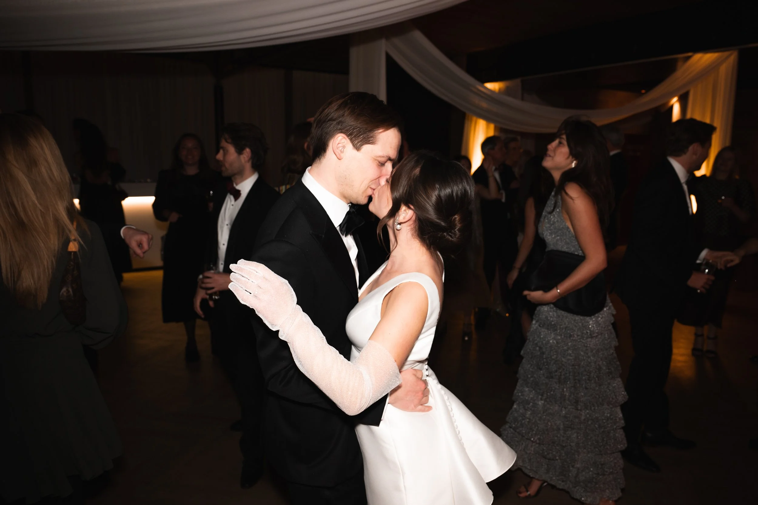 A young couple in formal attire sharing a dance at a wedding reception, surrounded by other guests in elegant clothing.