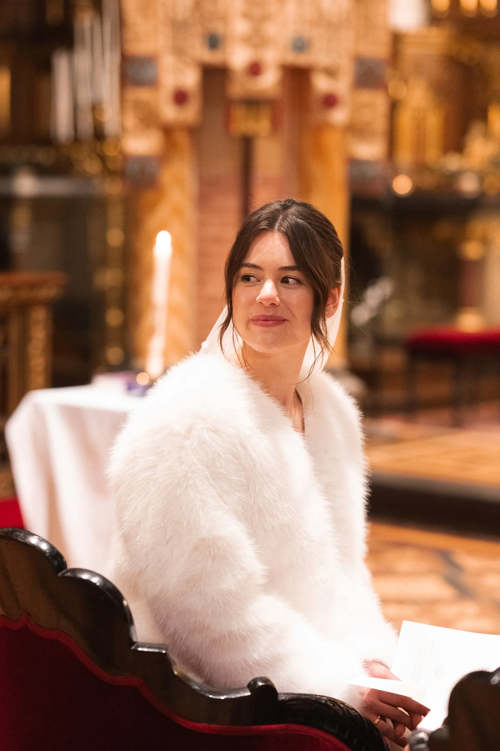 A woman with dark hair in an updo, wearing a white fluffy coat, sitting in a decorated room with brick walls and wooden accents, holding a piece of paper and smiling softly.