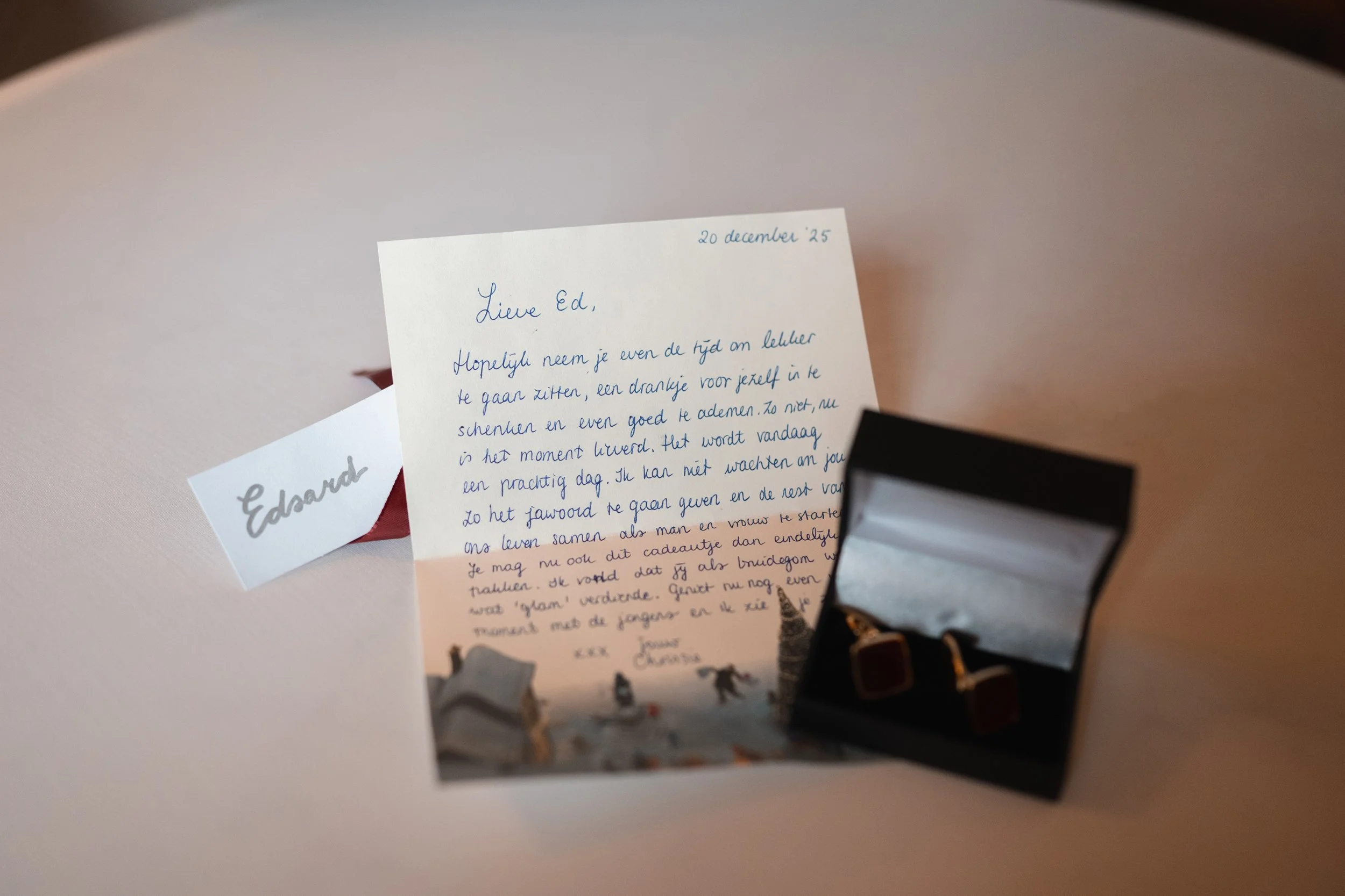 A handwritten letter dated December 20, 2025, is placed on a table. The letter is addressed to Ed and written in blue ink, with a small holiday-themed illustration at the bottom. Next to it is a white card with the name 'Edward' written in gray ink. 