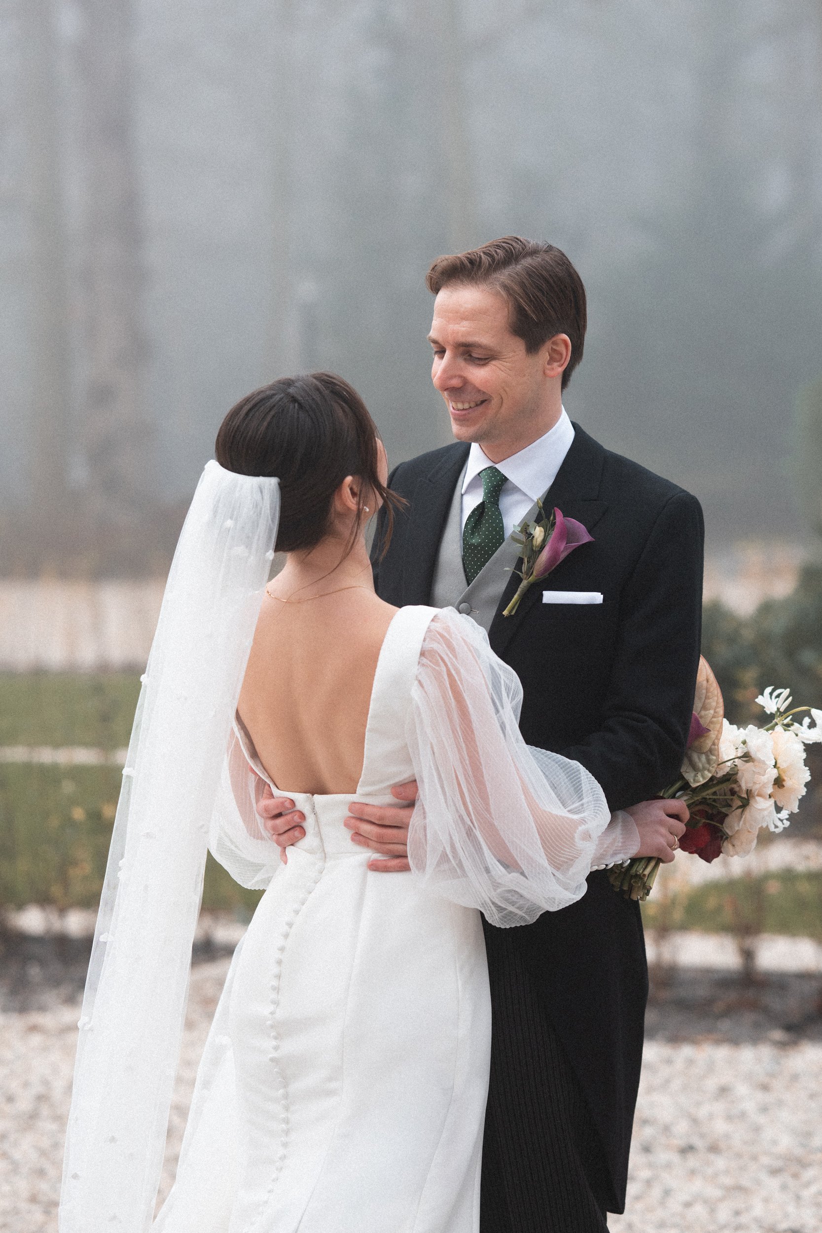 A bride and groom share a moment outdoors on their wedding day, smiling at each other with the bride holding a bouquet of flowers.
