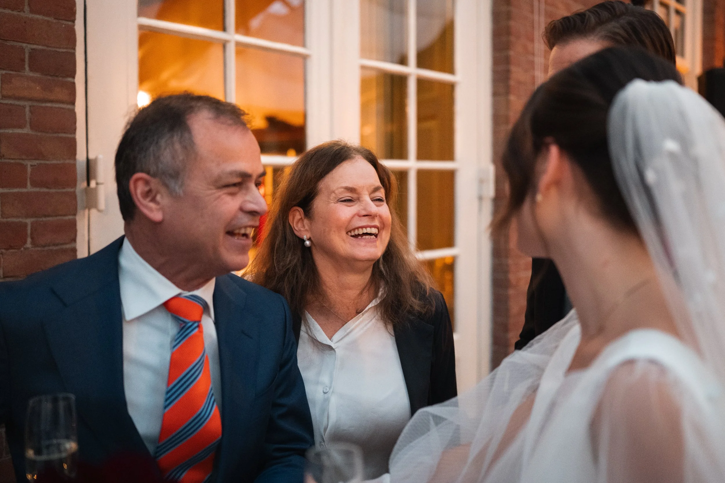 A joyful group of people laughing and talking at a wedding reception outside a building with brick walls and large windows.