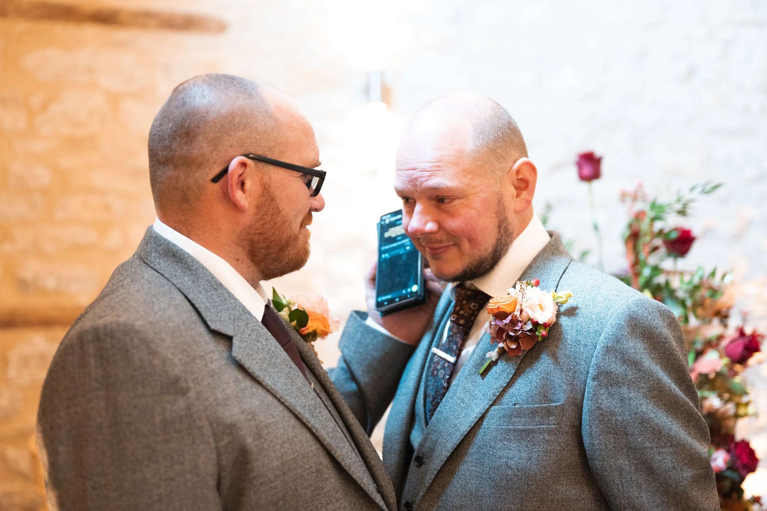 Two men in suits at a wedding, one holds a phone to the other's ear.