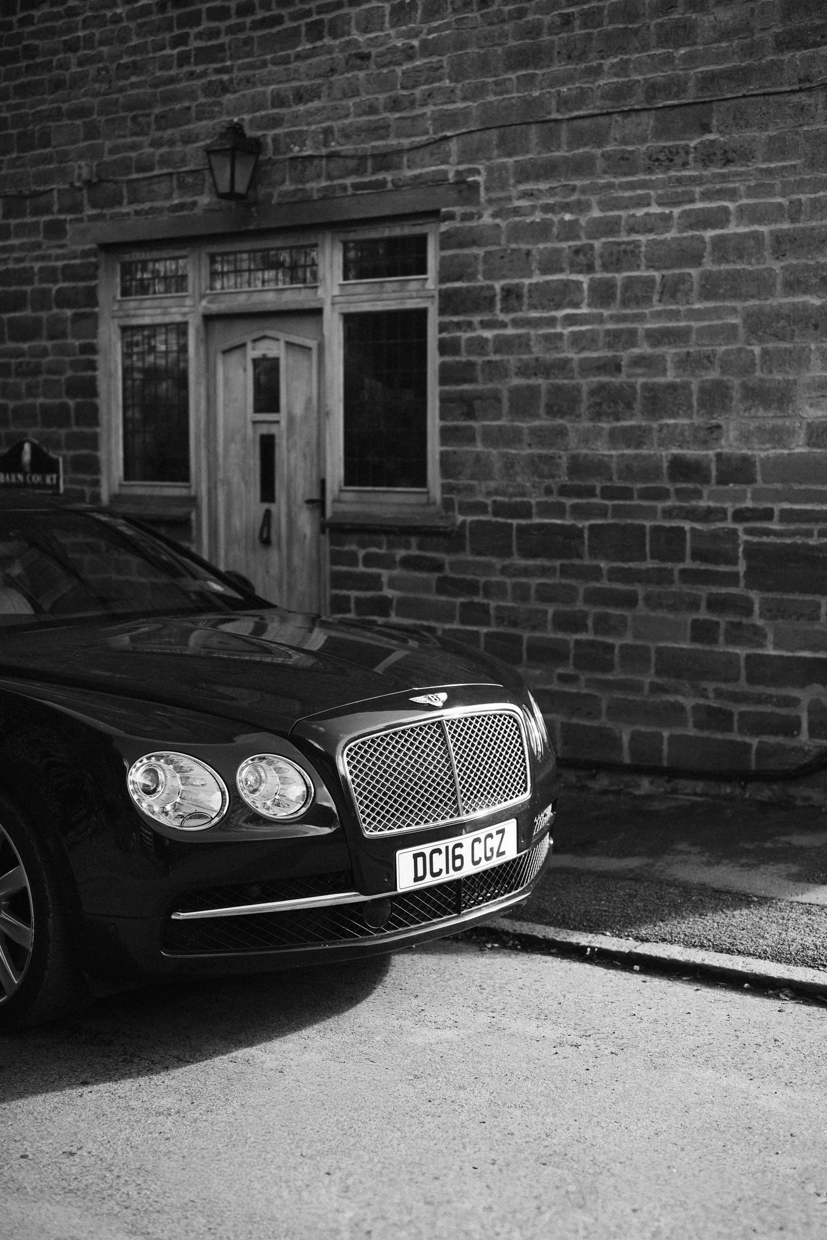Black luxury car parked on a residential street in front of a brick house with a wooden door and window panels, and a wall-mounted lantern light.
