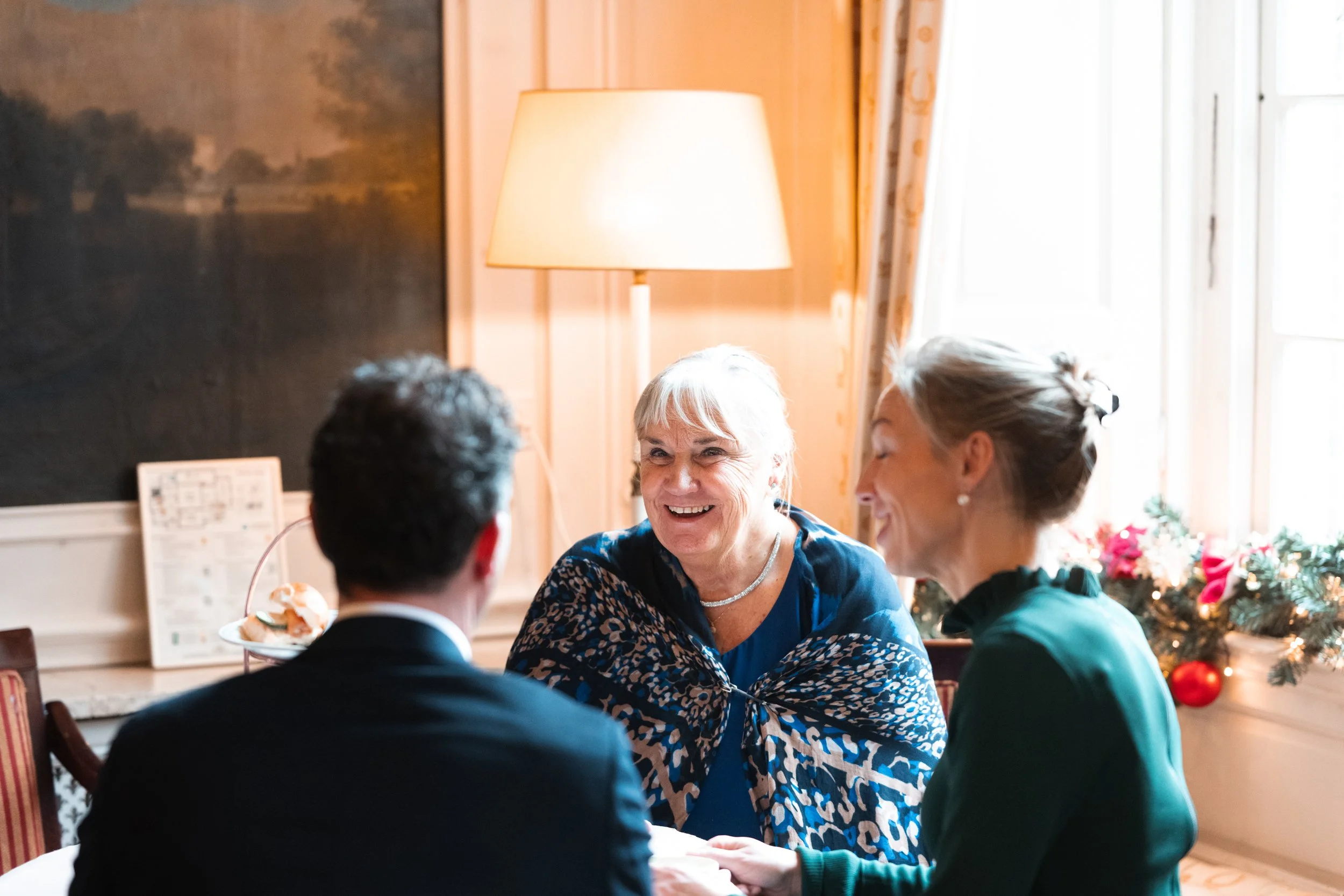 Three adults, two women and a man, are sitting at a table, smiling and holding hands in a cozy, decorated room with a window, a lamp, and Christmas decorations.