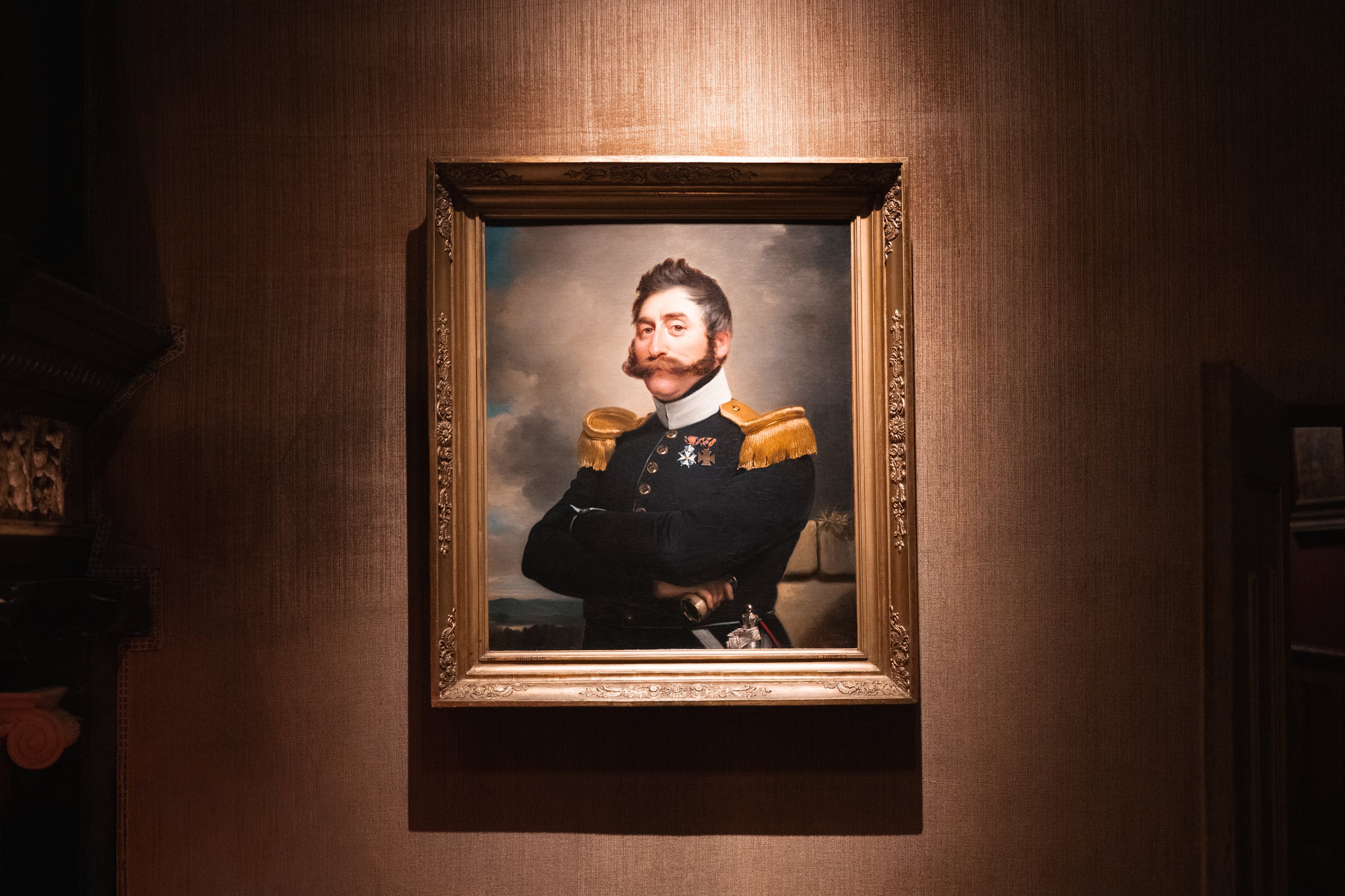 Portrait painting of a man in 19th-century military uniform with epaulets, crossed arms, and a mustache, set in a decorative frame on a textured wall.