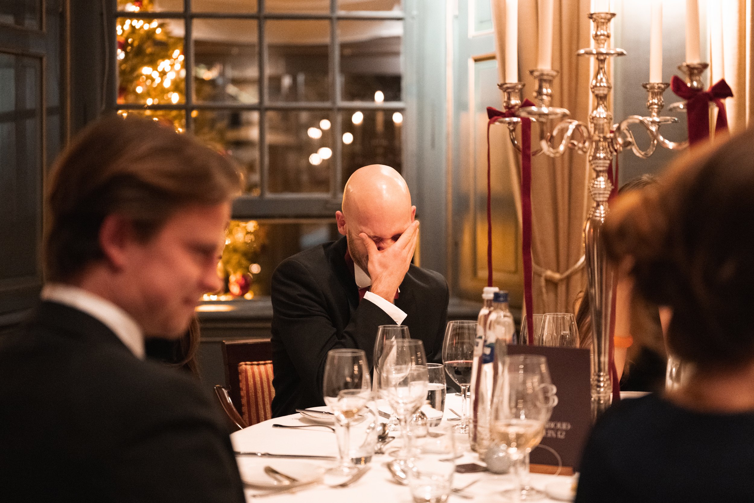 A man in a suit with his hand covering his face sitting at a dinner table during a holiday celebration, surrounded by wine glasses and candles, with a Christmas tree in the background.