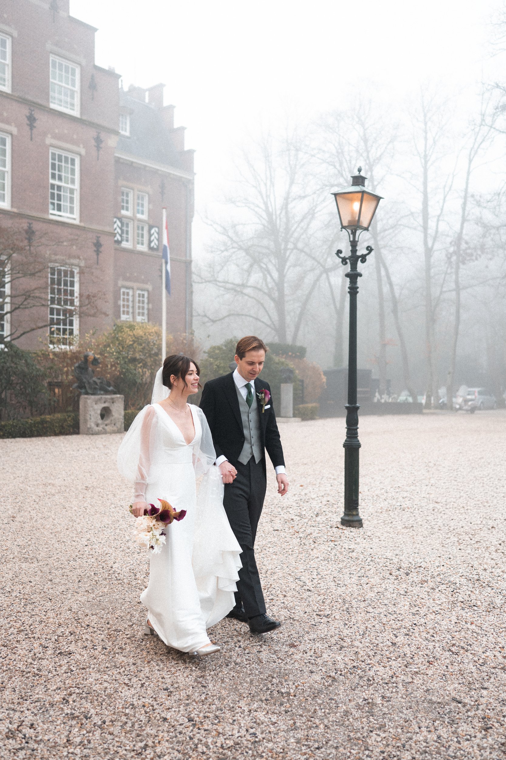 A bride and groom walking together outdoors on a foggy day, holding hands, with a historic building and a lamp post in the background.