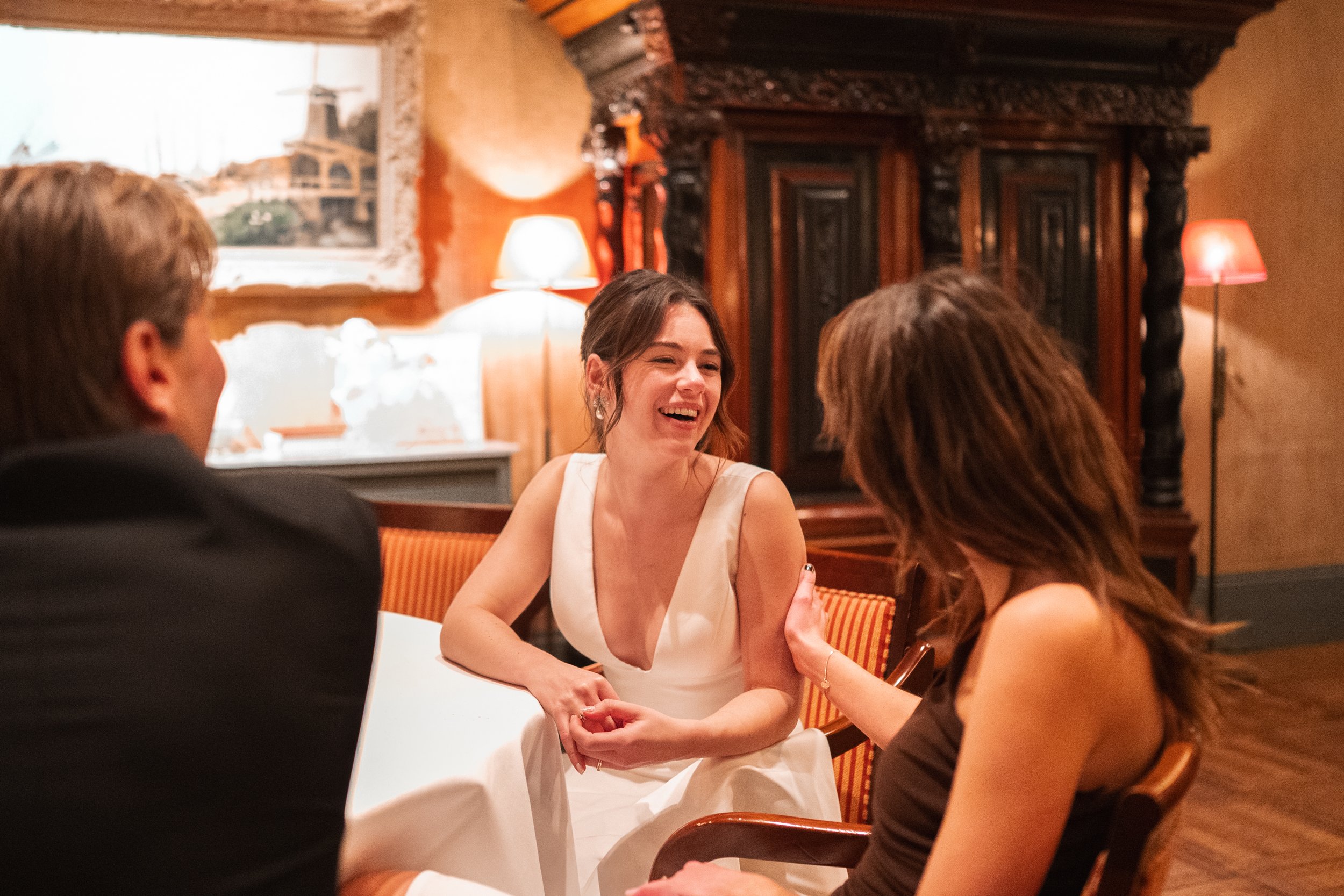 A woman in a white dress is laughing while talking to two people, a man and a woman, in a warmly lit, cozy room with wooden walls and vintage furniture.
