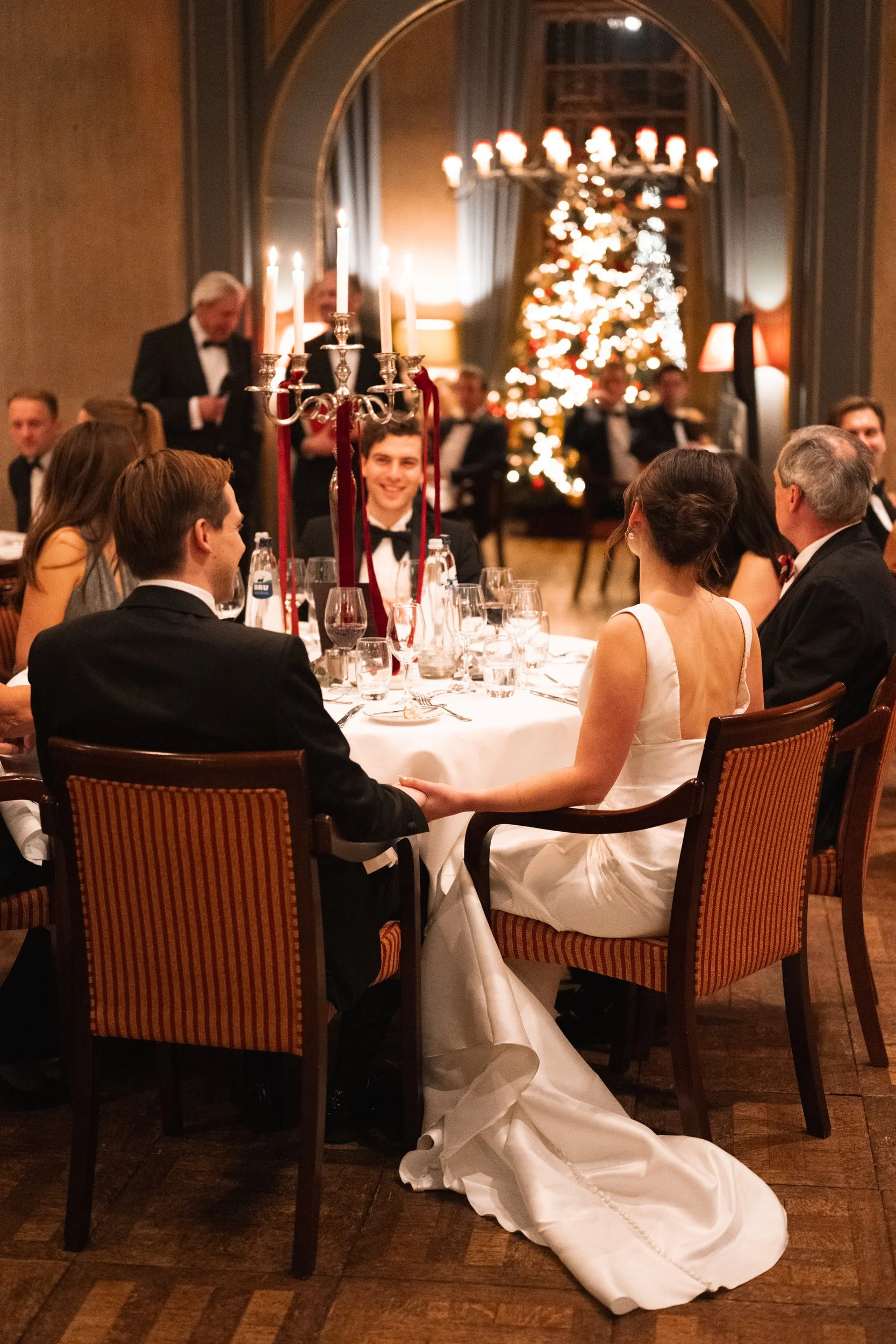 A couple dressed in formal wedding attire sitting at a round table during a holiday celebration, with a Christmas tree in the background and elegant decor.