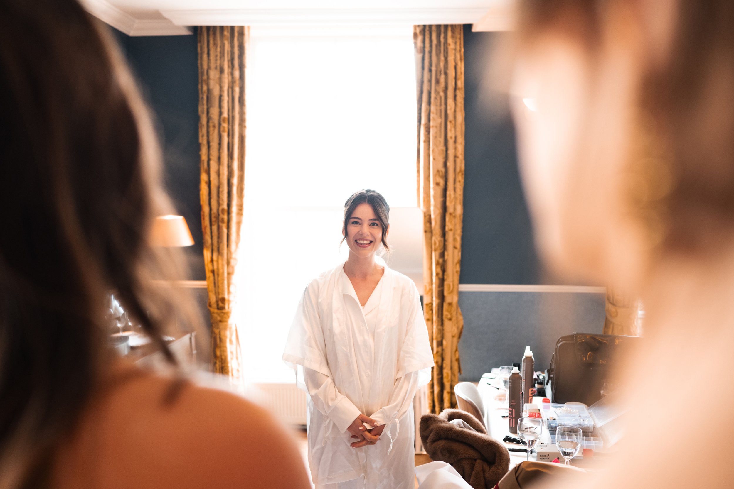 A young woman smiling in a white gown, surrounded by others, standing in a room with gold curtains and a large window in the background.