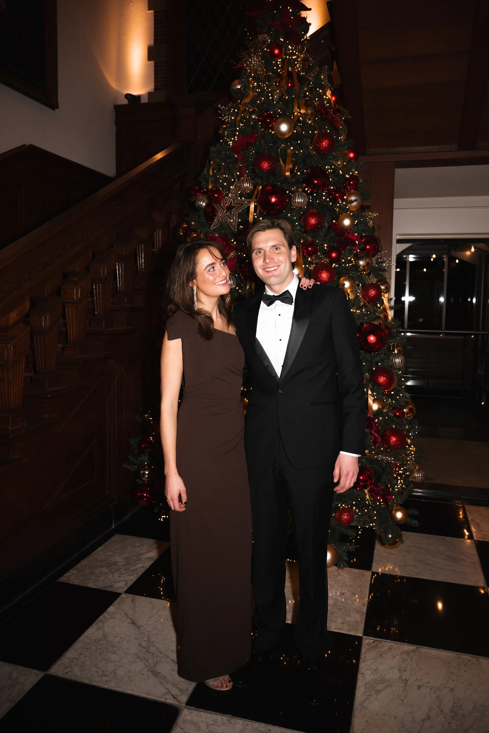 A couple dressed in formal attire standing in front of a decorated Christmas tree indoors. The woman is wearing a brown gown and the man is in a black tuxedo with a bow tie. The Christmas tree has red, gold, and silver ornaments with lights, and the 