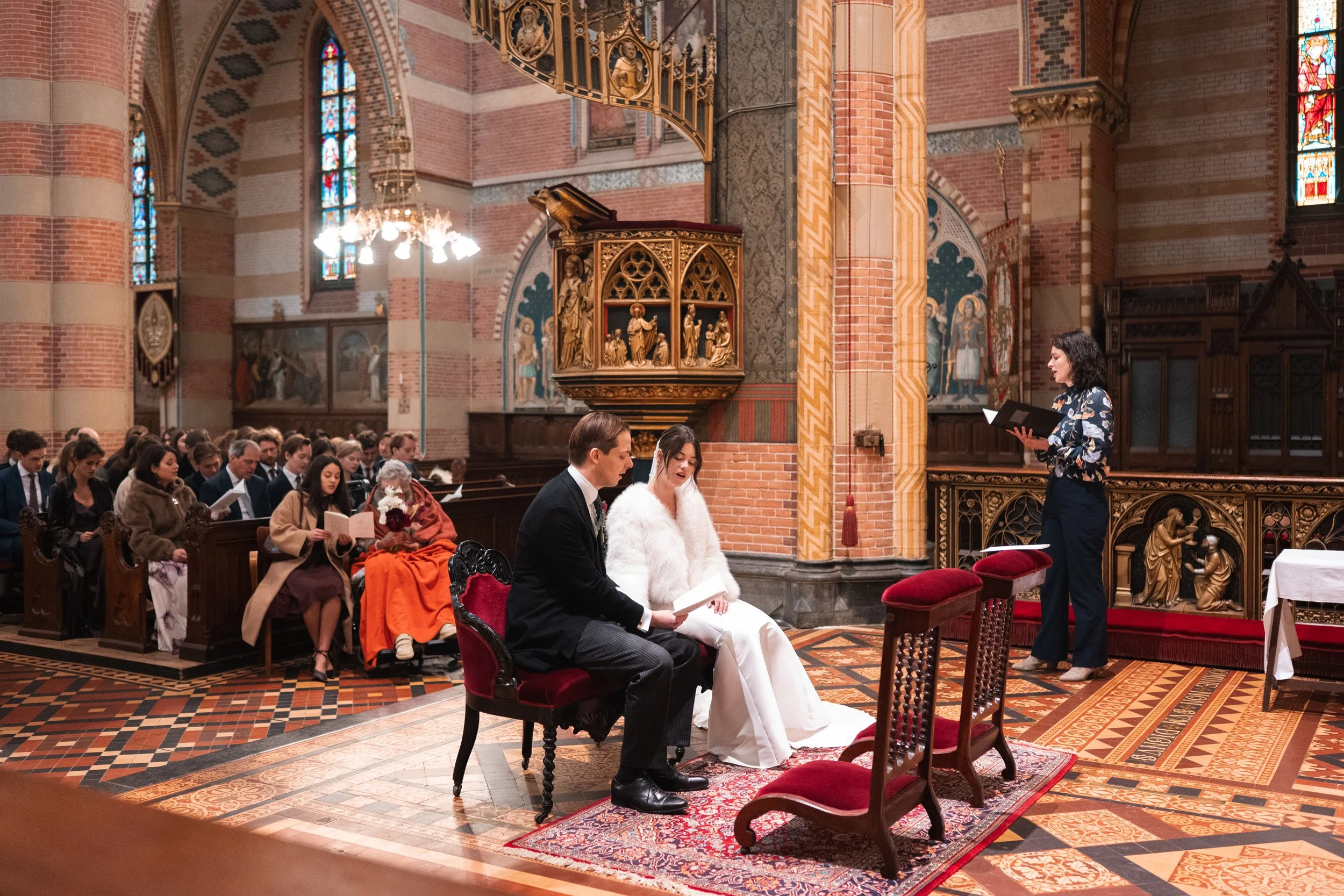 A wedding ceremony inside a church with a couple sitting in front of an officiant. The bride wears a white dress and faux fur coat, while the groom wears a black suit. Several guests sit on pews watching the ceremony.