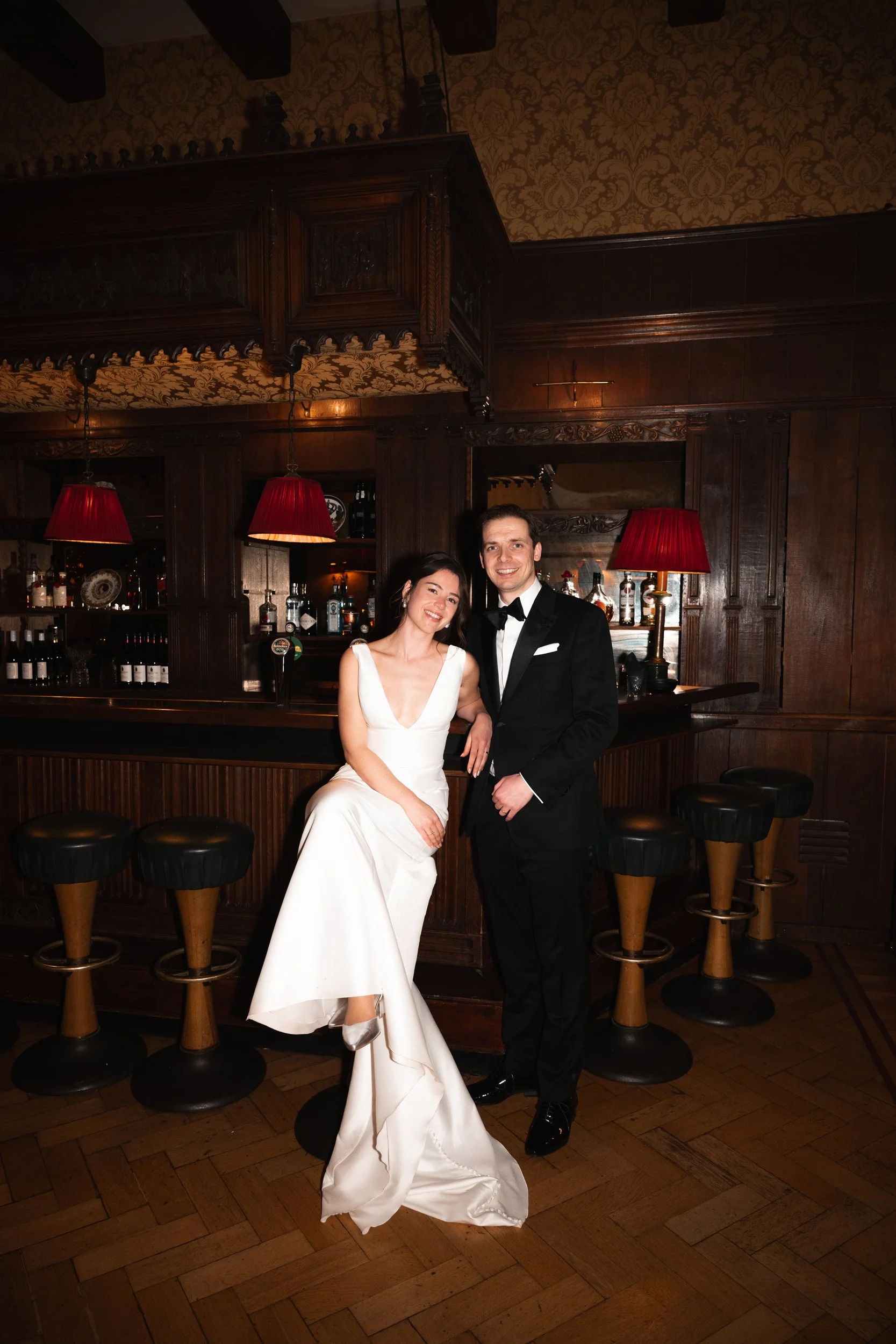 A bride and groom in formal wedding attire posing inside a dimly lit bar with dark wood paneling, bar stools, and red lampshades.