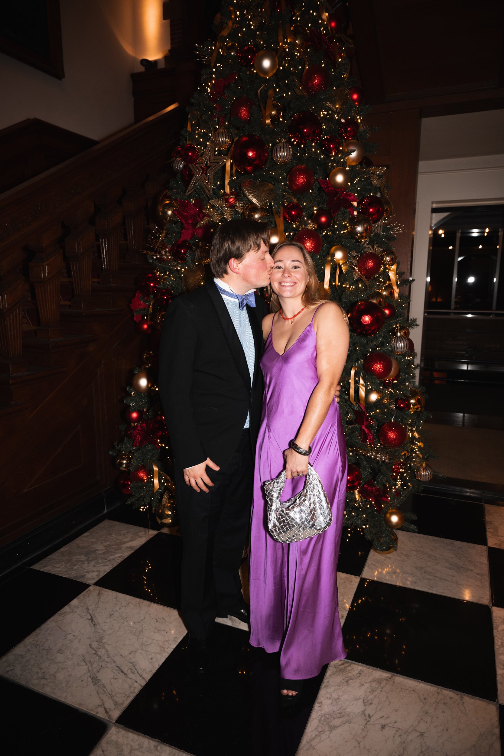 A man in a black suit and a woman in a purple dress standing in front of a decorated Christmas tree, with the man kissing the woman's cheek. The woman is holding a silver clutch purse and smiling.