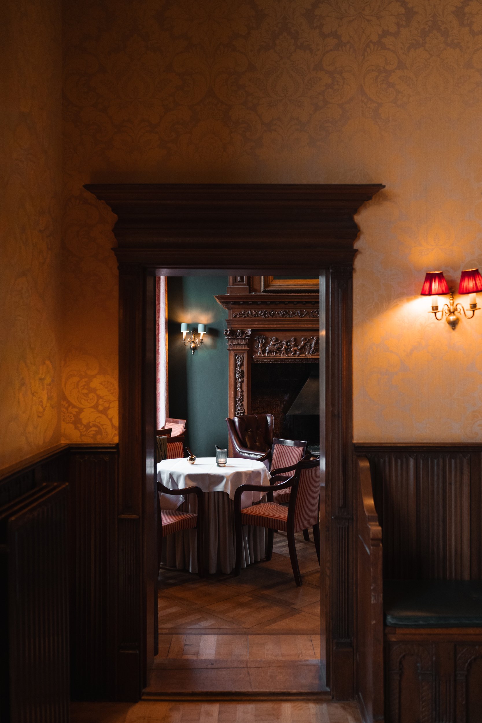 Elegant dining room with a round table covered with a white tablecloth, surrounded by red and wooden chairs, seen through a dark wooden doorway with ornate trim, with warm lighting and a fireplace in the background.