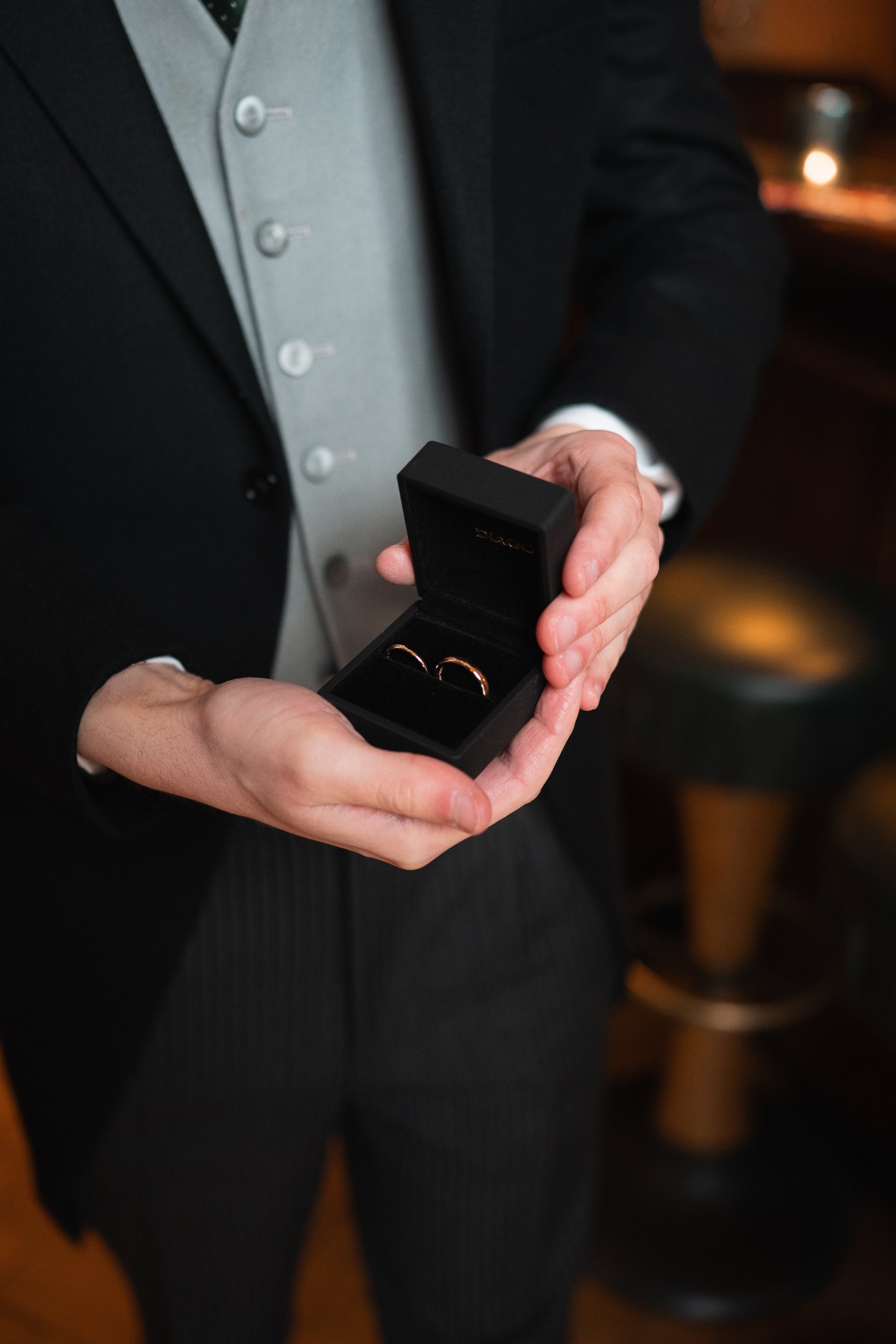 A person dressed in formal attire holding a black jewelry box with two gold wedding rings inside.