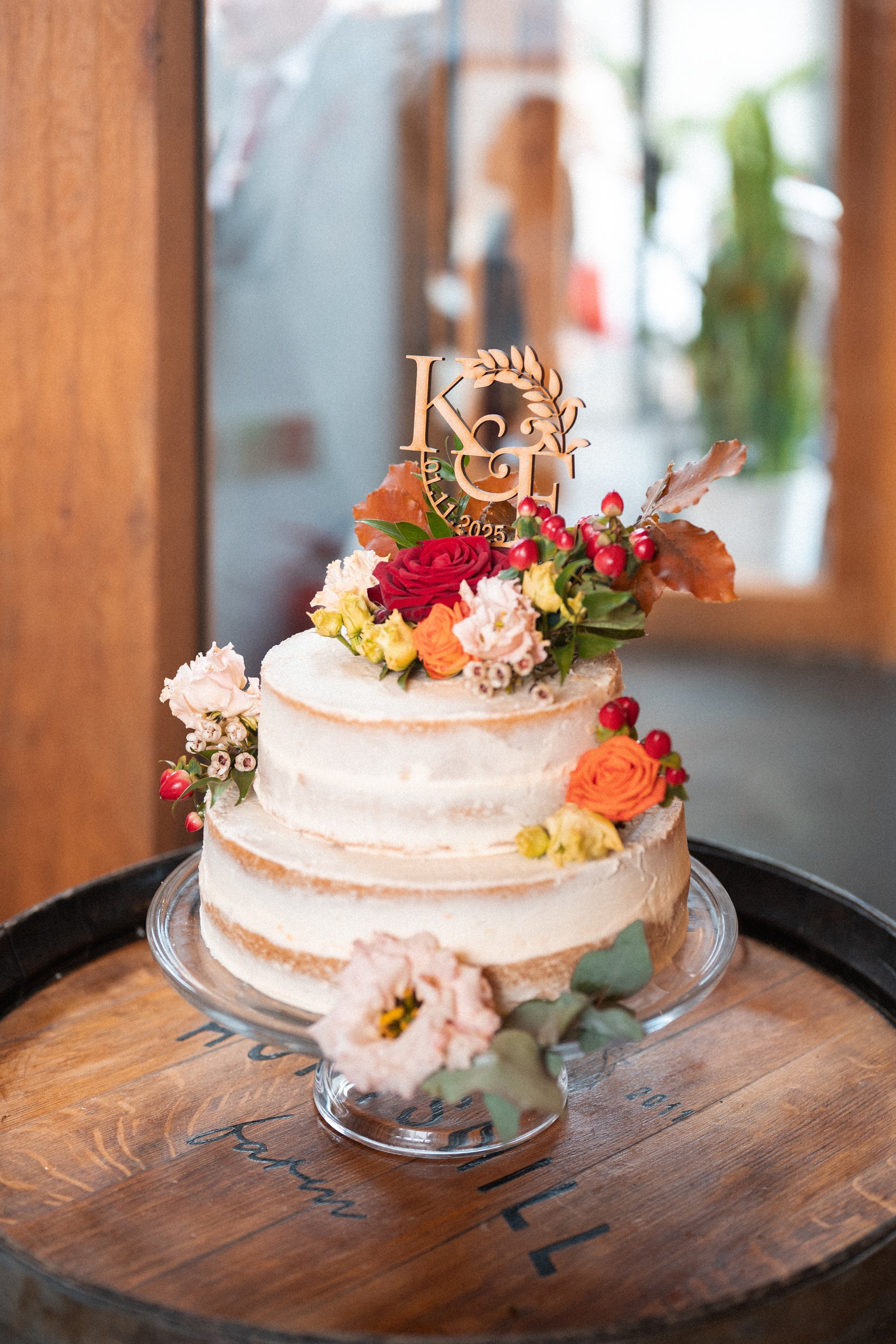 A wedding or celebration cake decorated with fresh flowers and a wooden topper with initials 'K & E' and the date 2025, placed on a wooden barrel.
