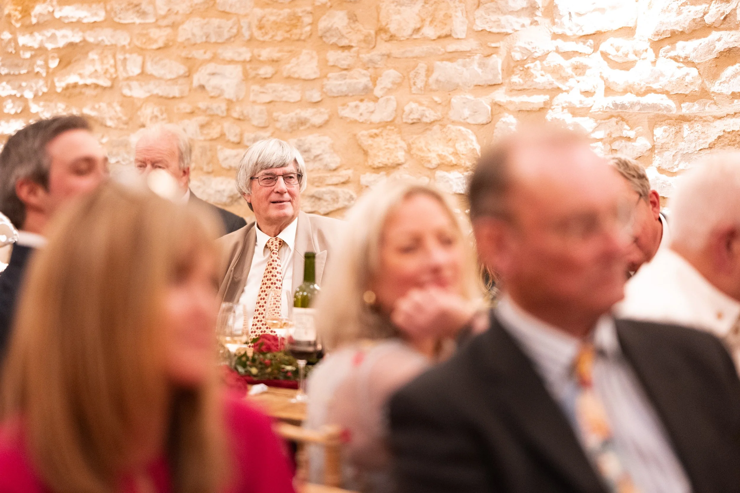 People seated at a dinner event in front of a stone wall, including an older man with gray hair and glasses wearing a tan suit and patterned tie, among other attendees.