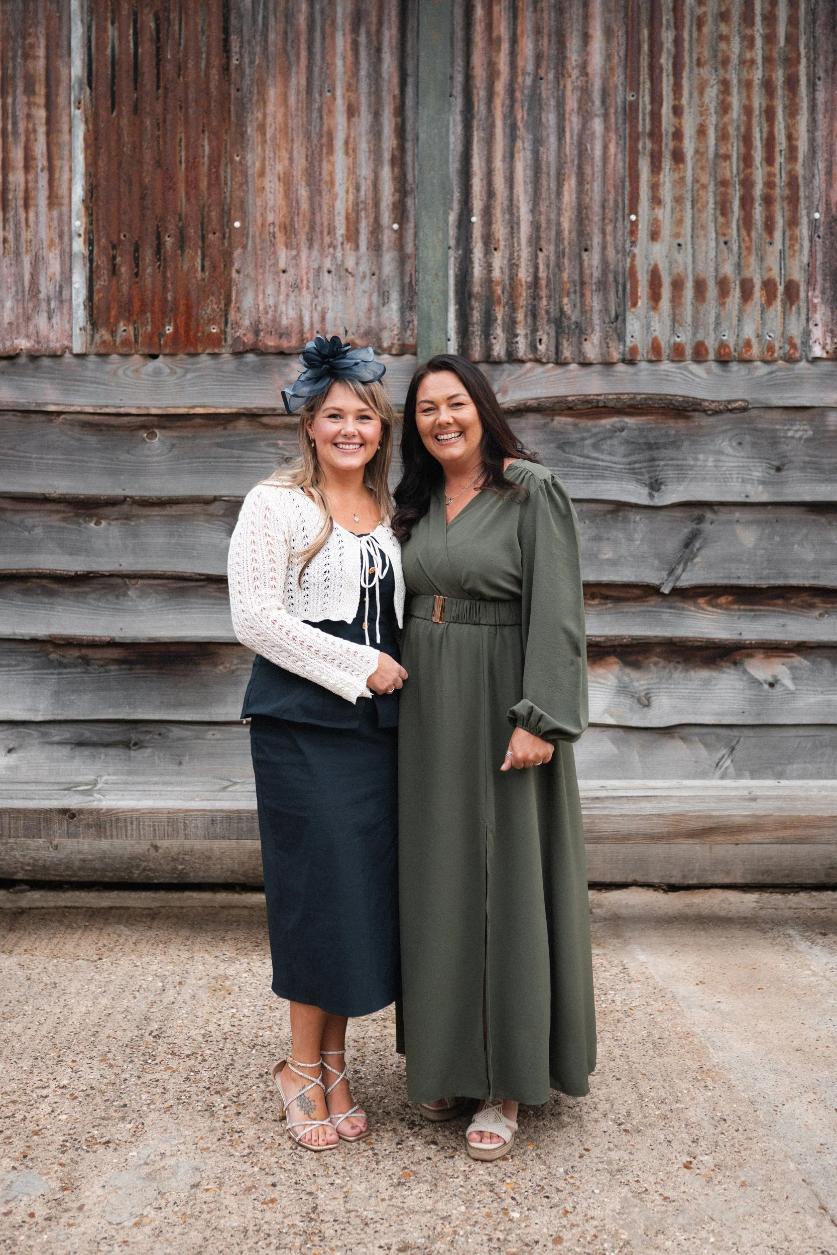 Two women smiling and standing close together in front of a rustic wooden and metal wall.