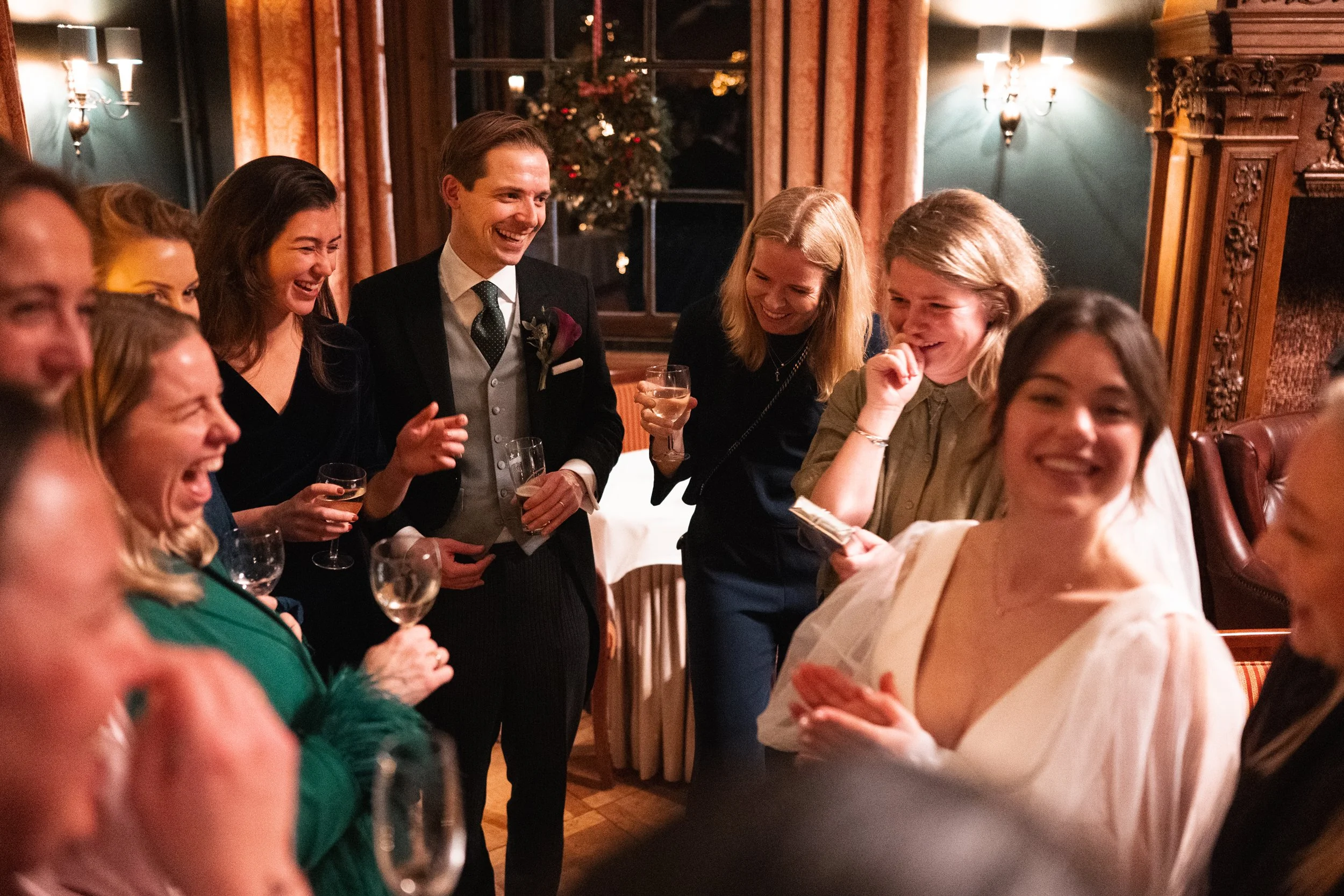 Group of people at a celebration or party, laughing and holding drinks in a warmly decorated room.