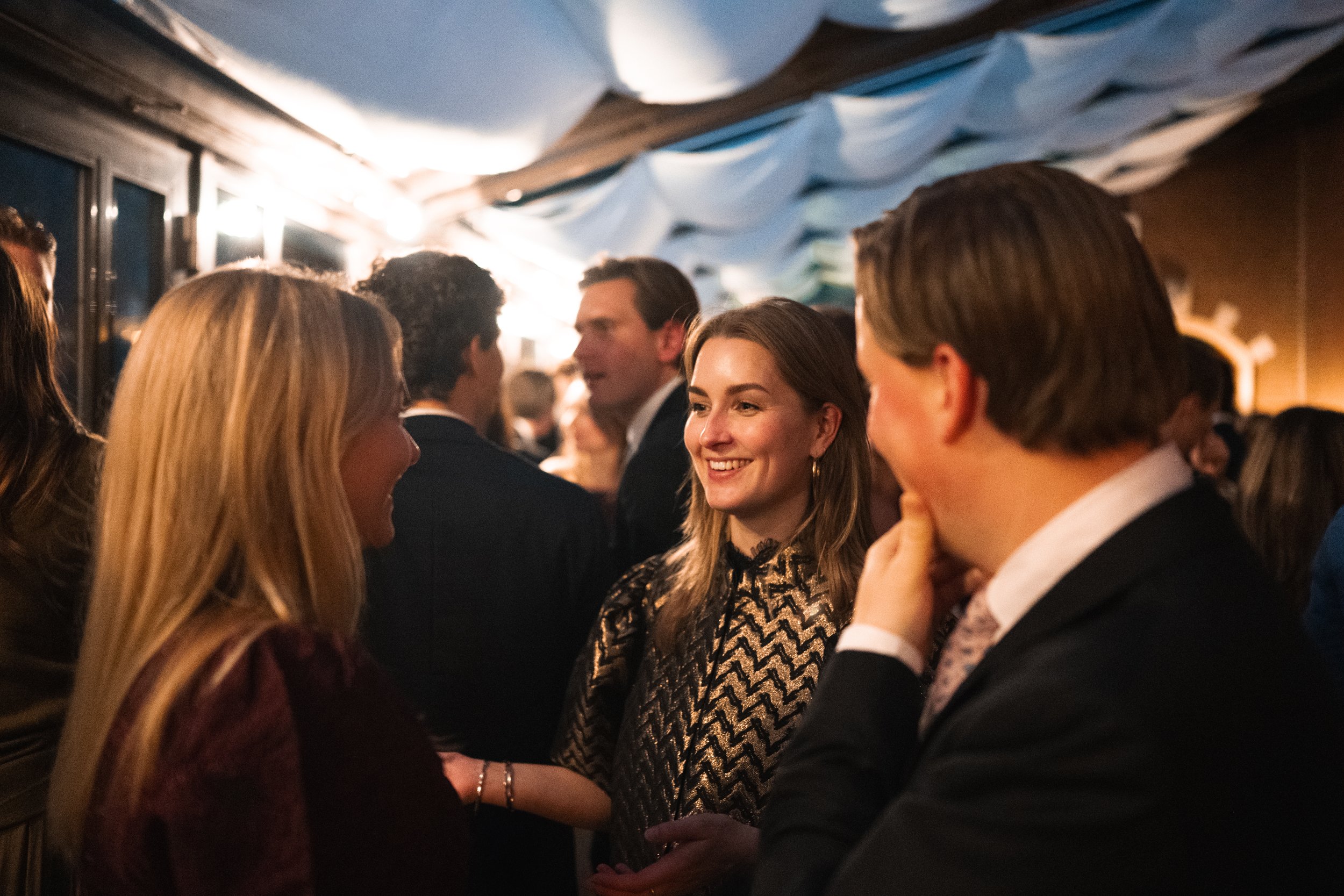 A group of people socializing at an indoor event, including a woman smiling and talking with a man, with other guests in the background.